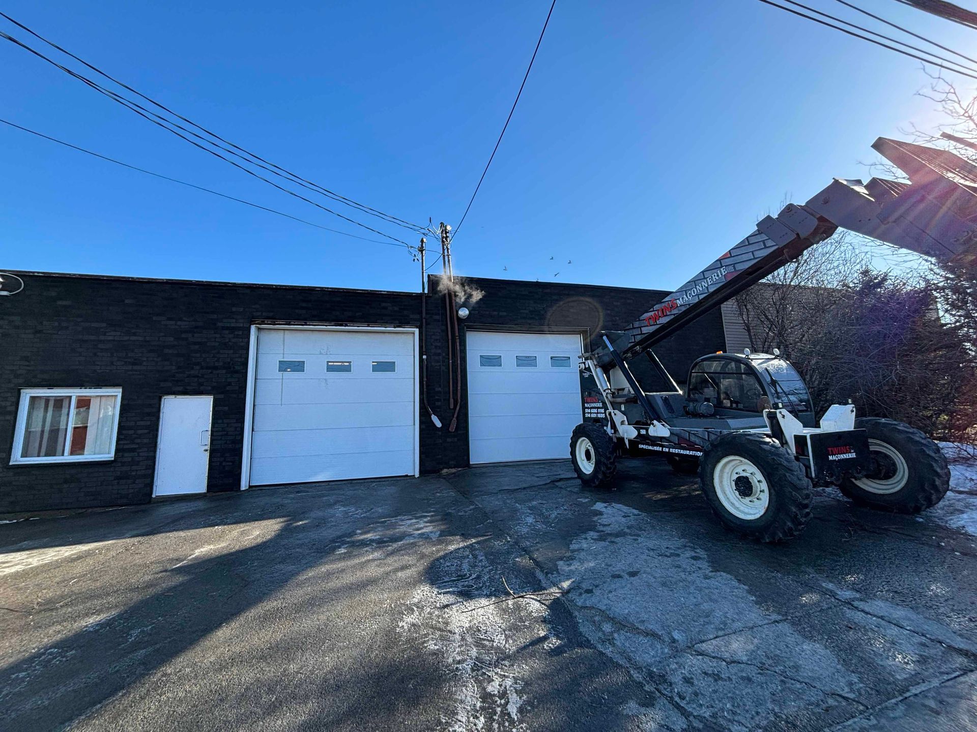 Bâtiment noir avec portes de garage blanches et chariot télescopique par une journée ensoleillée et enneigée.