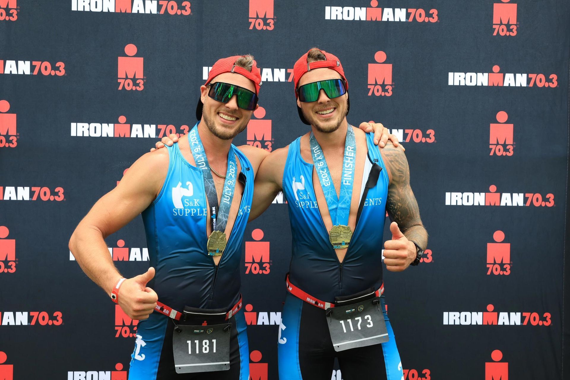 Deux athlètes en tenue de course, bras dessus bras dessous, posent après un Ironman 70.3. Ils portent des médailles et des lunettes de soleil.