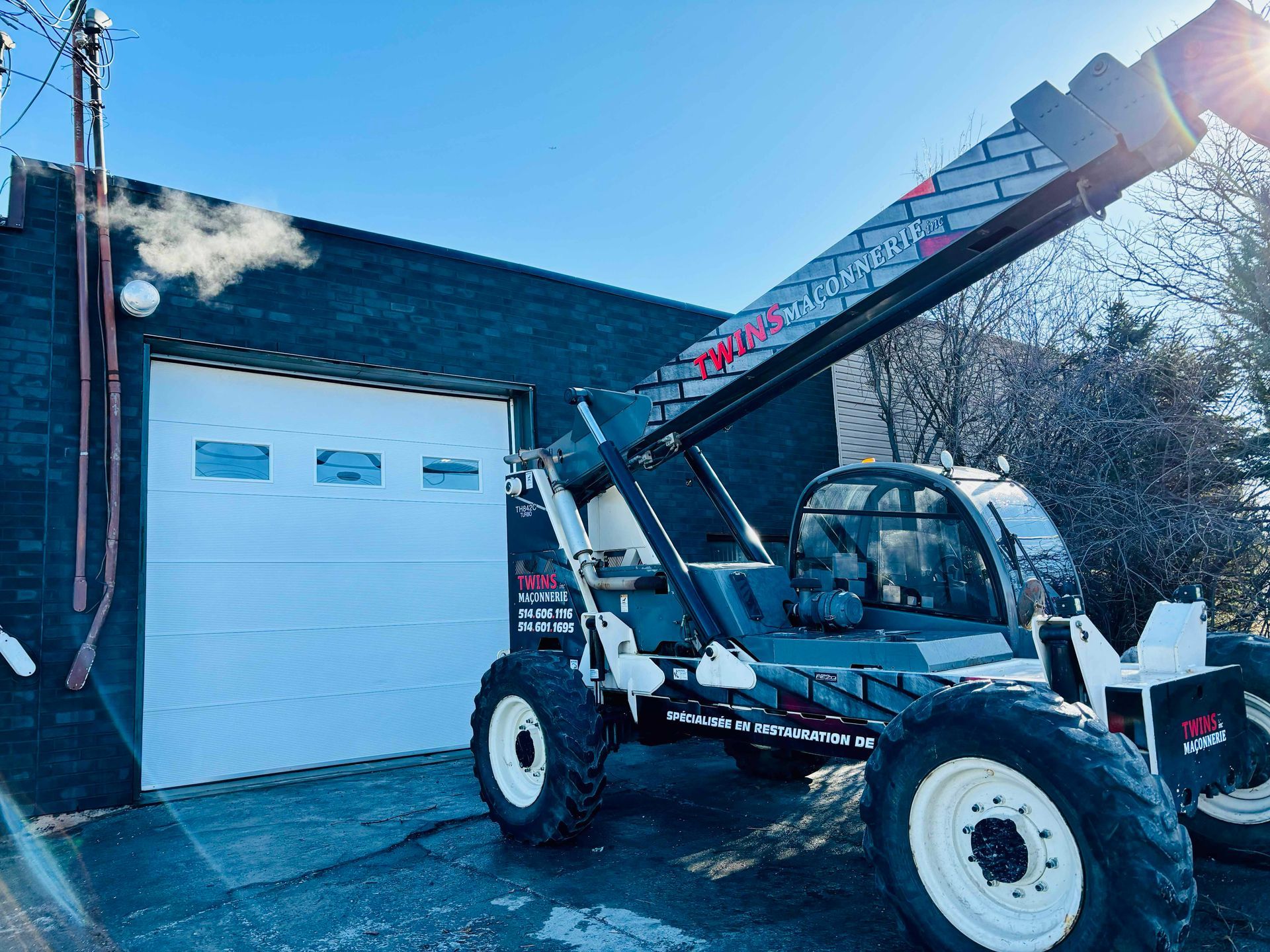 La flèche d'un chariot télescopique surplombe un bâtiment avec une porte de garage blanche. Temps froid et vapeur.