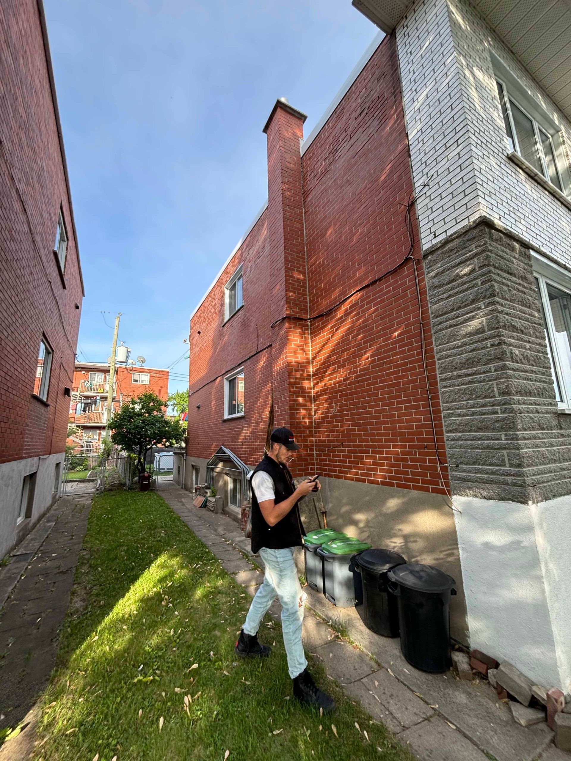 Un homme, tenant un téléphone, se trouve près d'un immeuble en briques. L'arrière-plan est une ruelle bordée d'herbe verte et de poubelles.