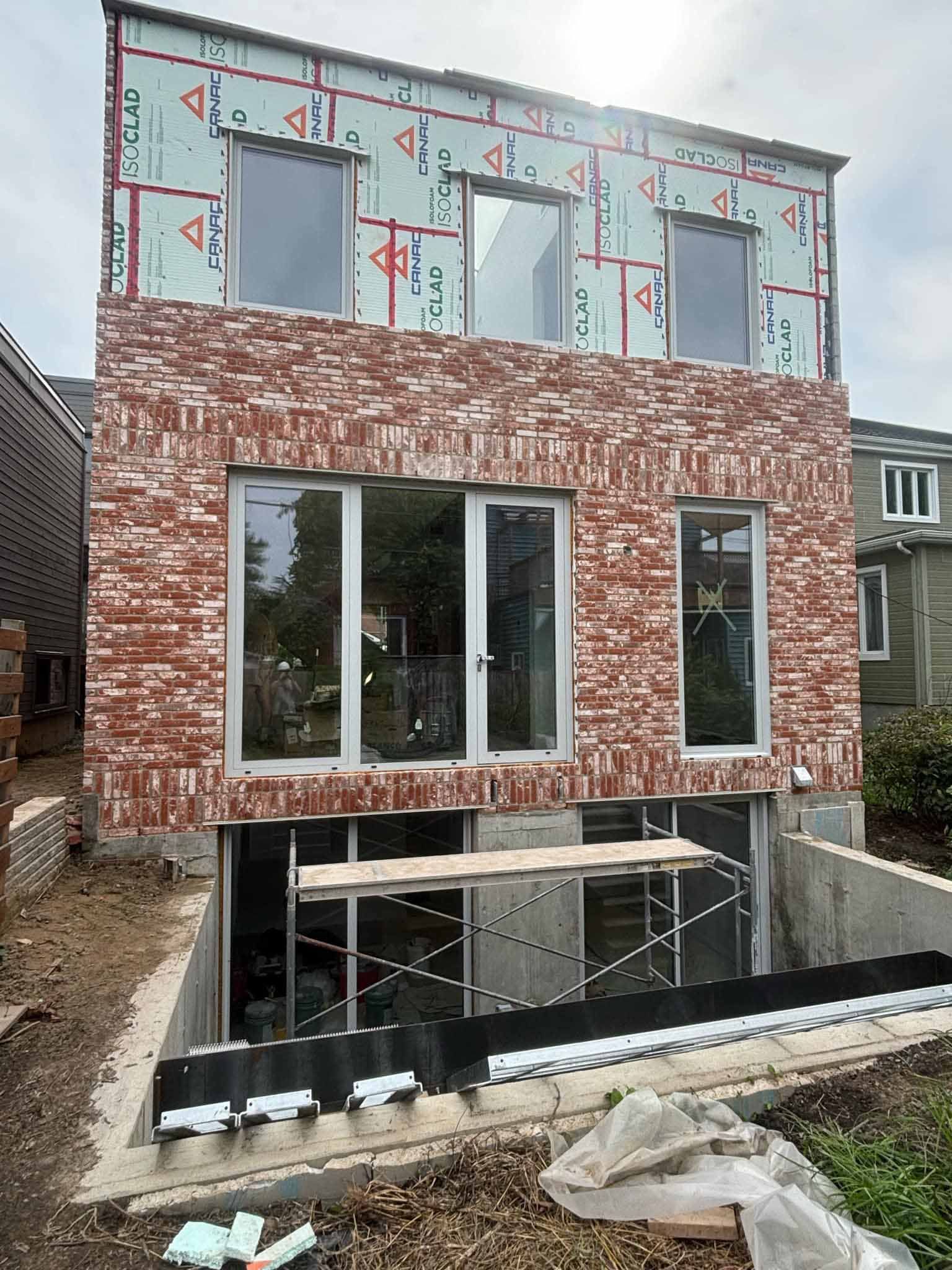 Bâtiment en briques en construction, fenêtres installées. Échafaudages visibles, sol en terre battue.