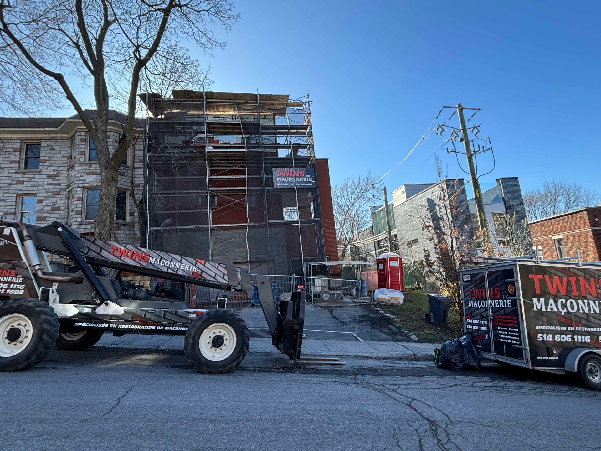 Chantier de construction avec échafaudages sur un bâtiment ; un chariot élévateur et une remorque portant la marque « Twin Maçons » à l'avant.