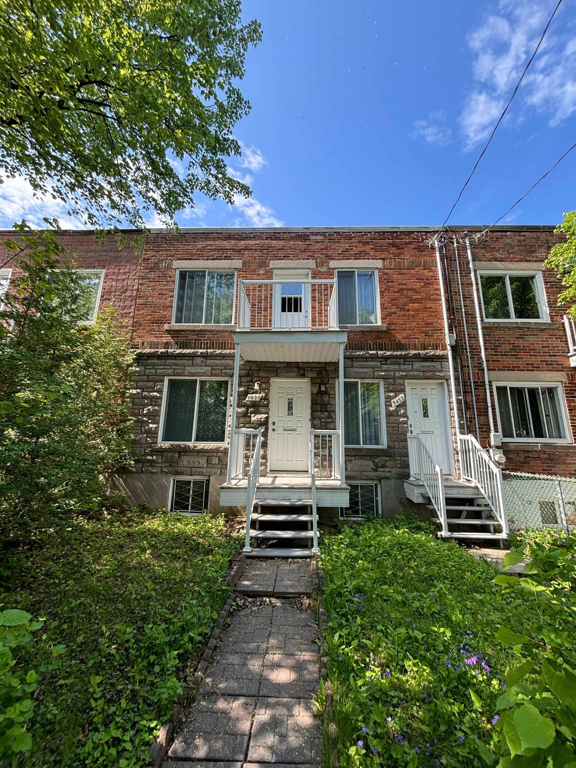 Immeuble de plusieurs logements en briques, avec portes et escaliers blancs, et végétation à l'avant.
