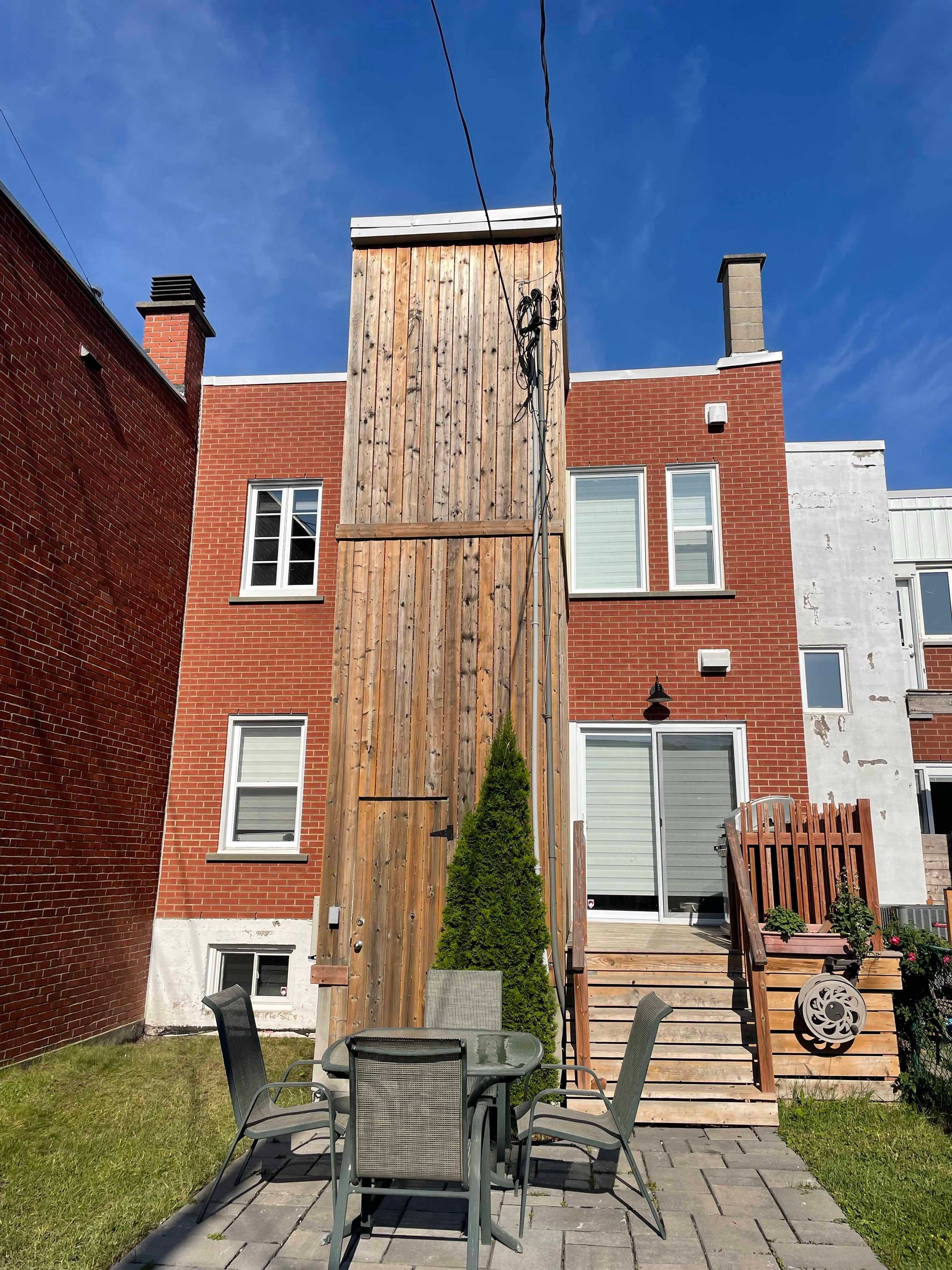 Jardin avec bâtiments en briques, structure en bois, mobilier de jardin et escaliers. Pelouse verte, ciel bleu.