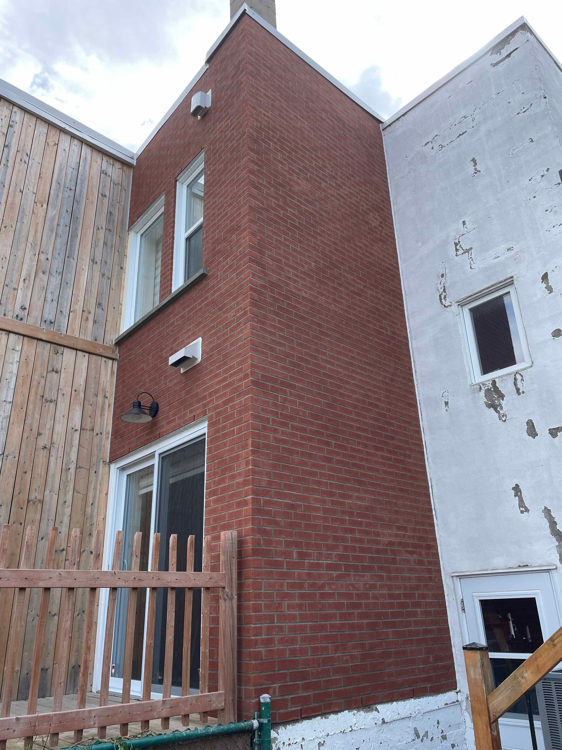 Bâtiment en briques rouges entre une clôture en bois et une structure en stuc blanc, avec des fenêtres et une cheminée.