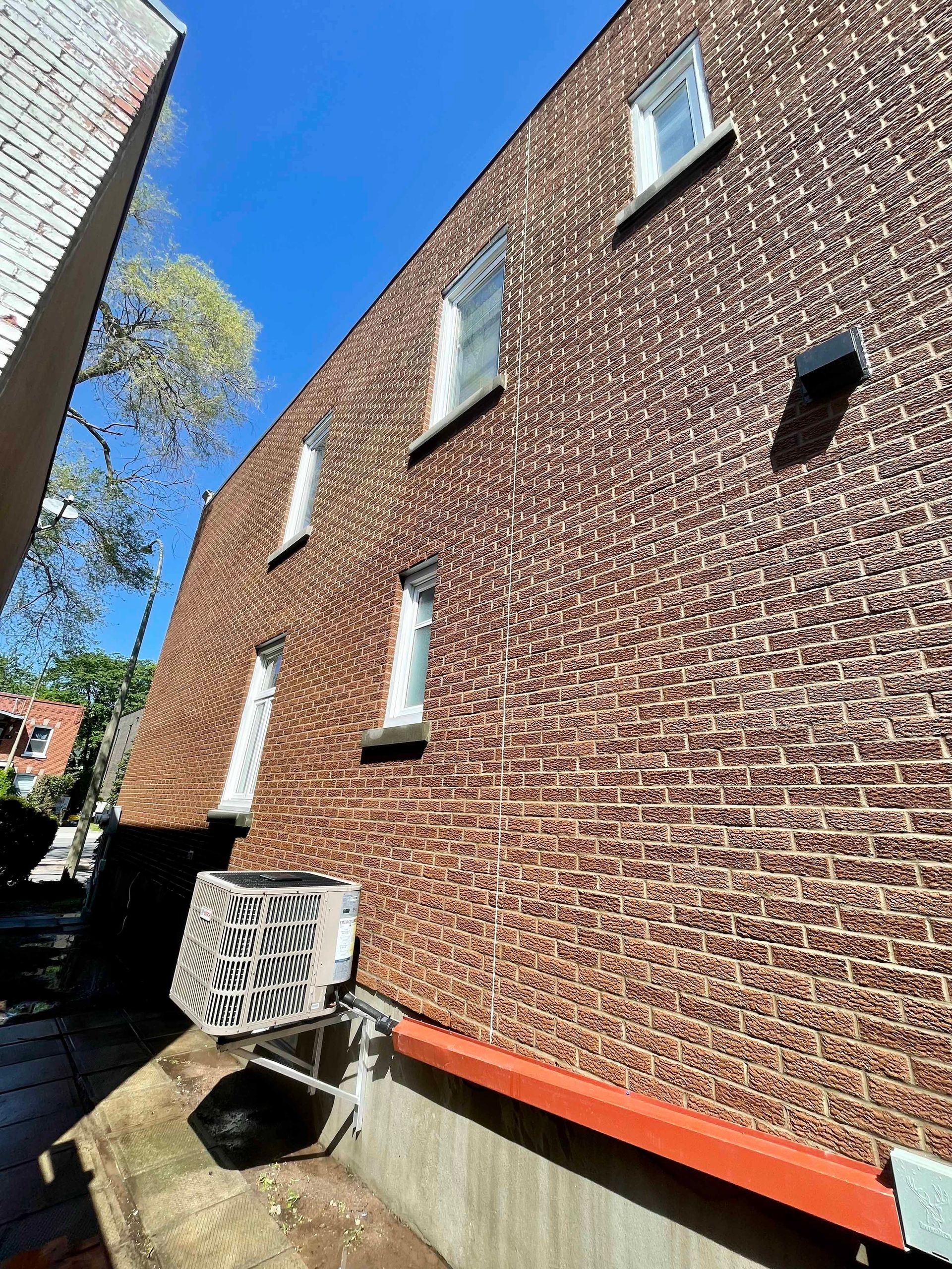 Bâtiment en briques avec fenêtres, climatiseur et poutre métallique orange à la base. Ciel bleu.
