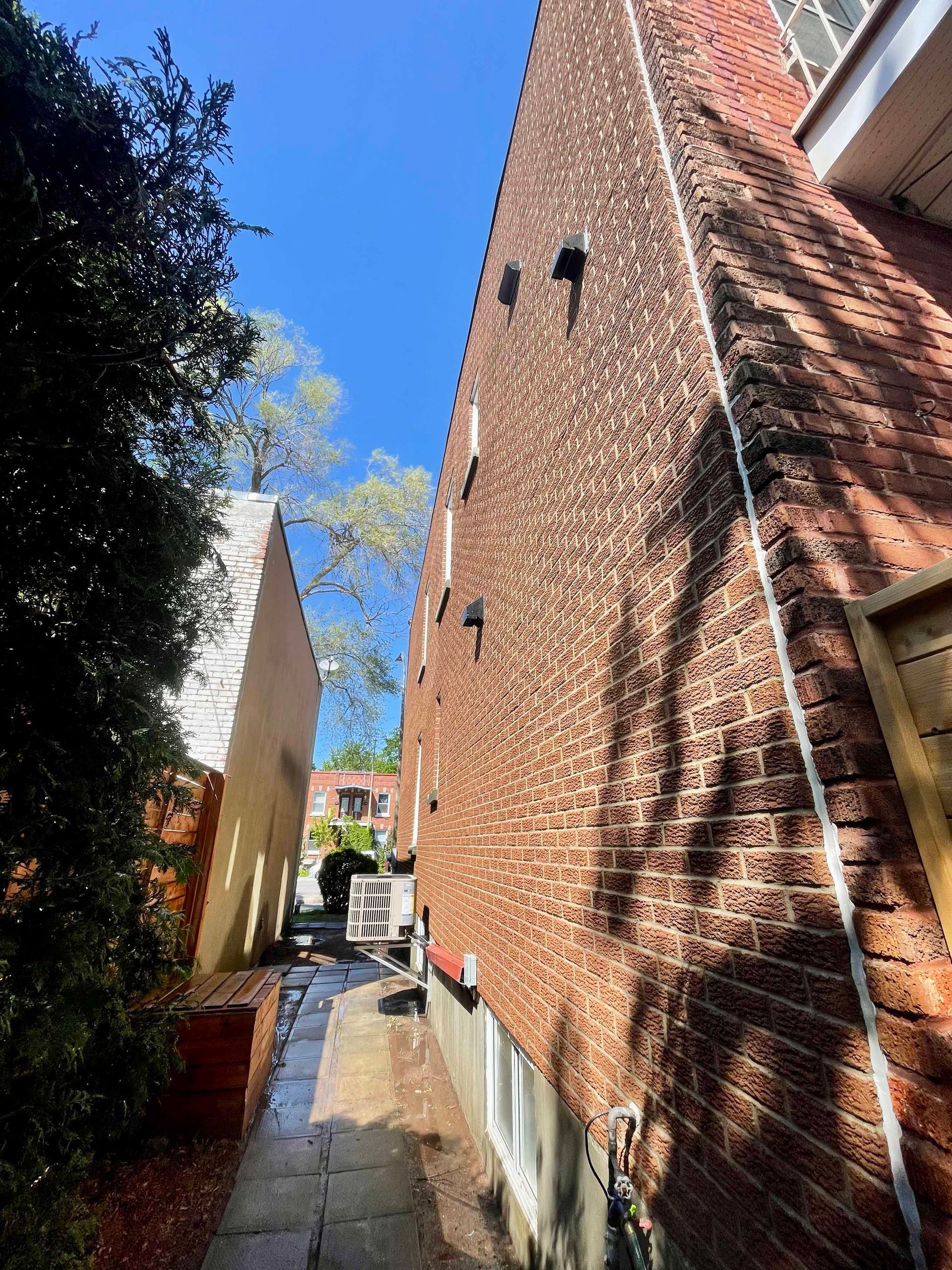 Façade d'un bâtiment en briques, jouxtant un mur blanc et une ruelle étroite par une journée ensoleillée.