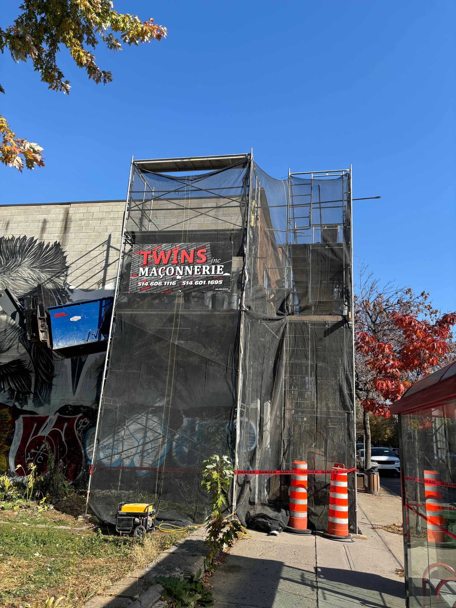 Façade du bâtiment recouverte d'échafaudages et de filets noirs, panneau « JIVE » au-dessus d'une allée clôturée.