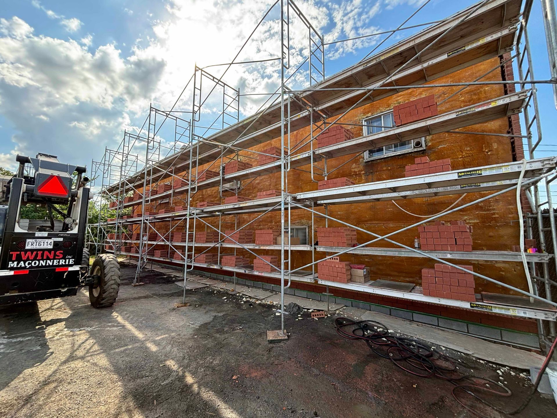 L'extérieur du bâtiment est en construction, avec des échafaudages et des briques rouges ; une chargeuse frontale est garée à proximité.