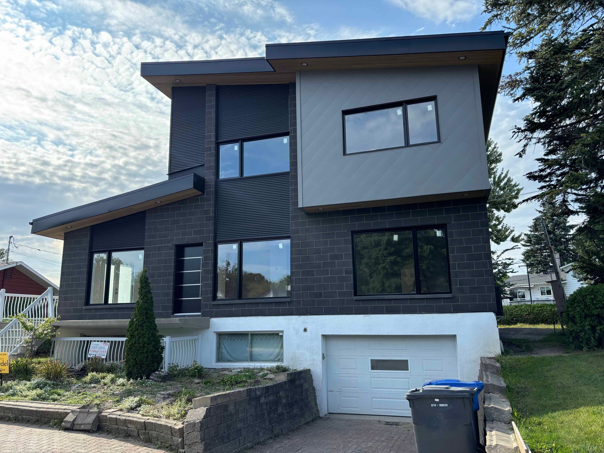 Maison moderne à deux étages avec briques foncées, bardage gris et porte de garage blanche. Poubelle de recyclage bleue devant.