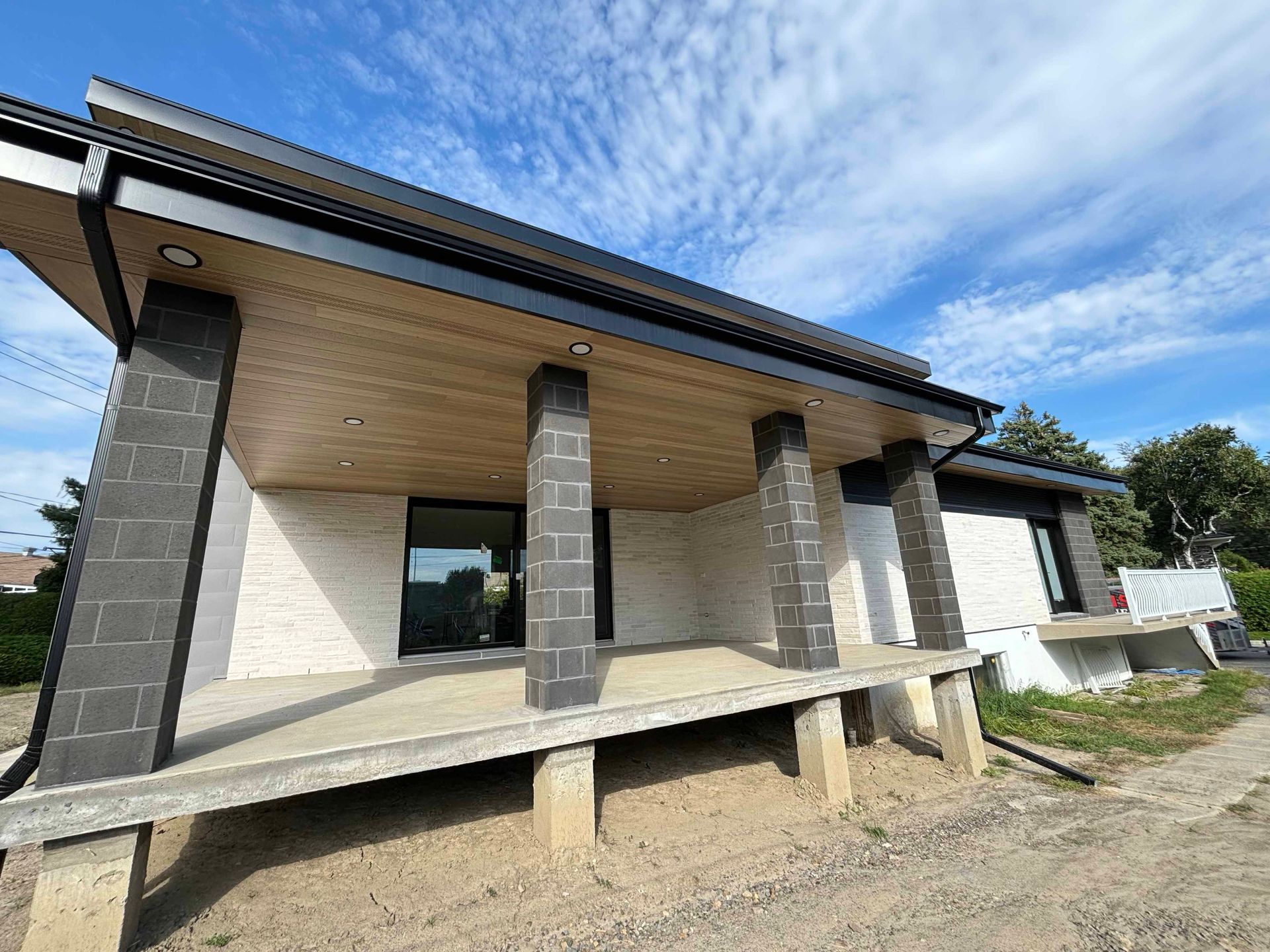 Maison moderne en construction avec un porche en béton et des piliers sombres, sur fond de ciel nuageux.