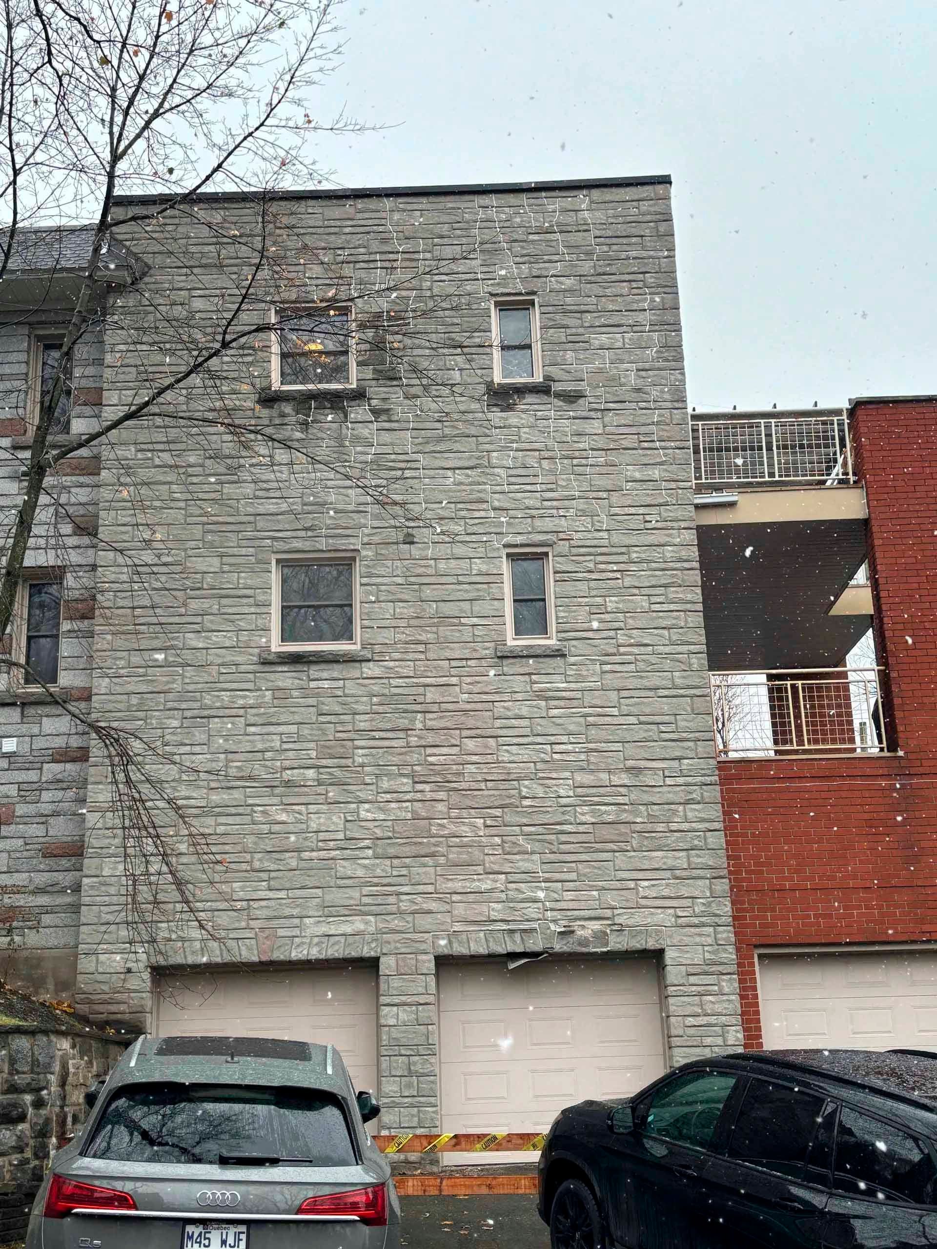 Bâtiment en pierre avec portes de garage, deux voitures garées devant et neige qui tombe.