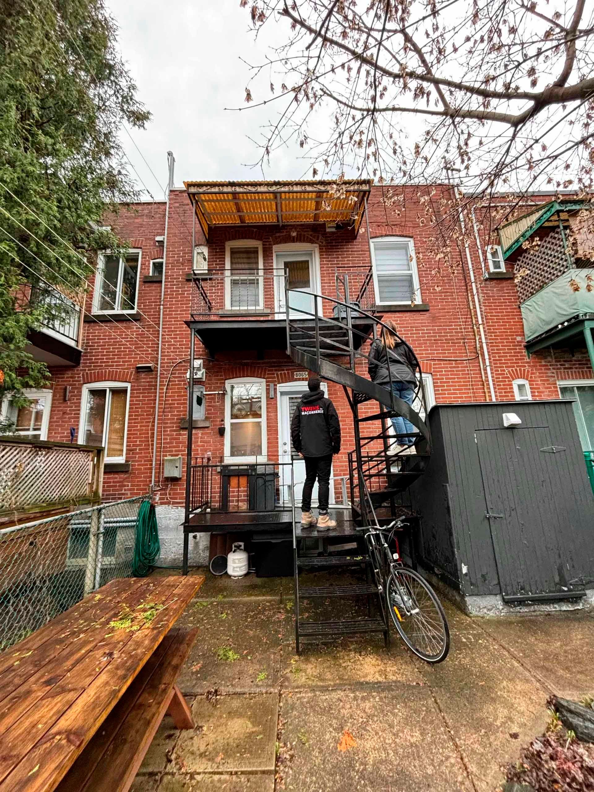 Extérieur d'un bâtiment en briques avec un escalier de secours métallique et deux personnes dessus. Un vélo est appuyé contre un mur à proximité.