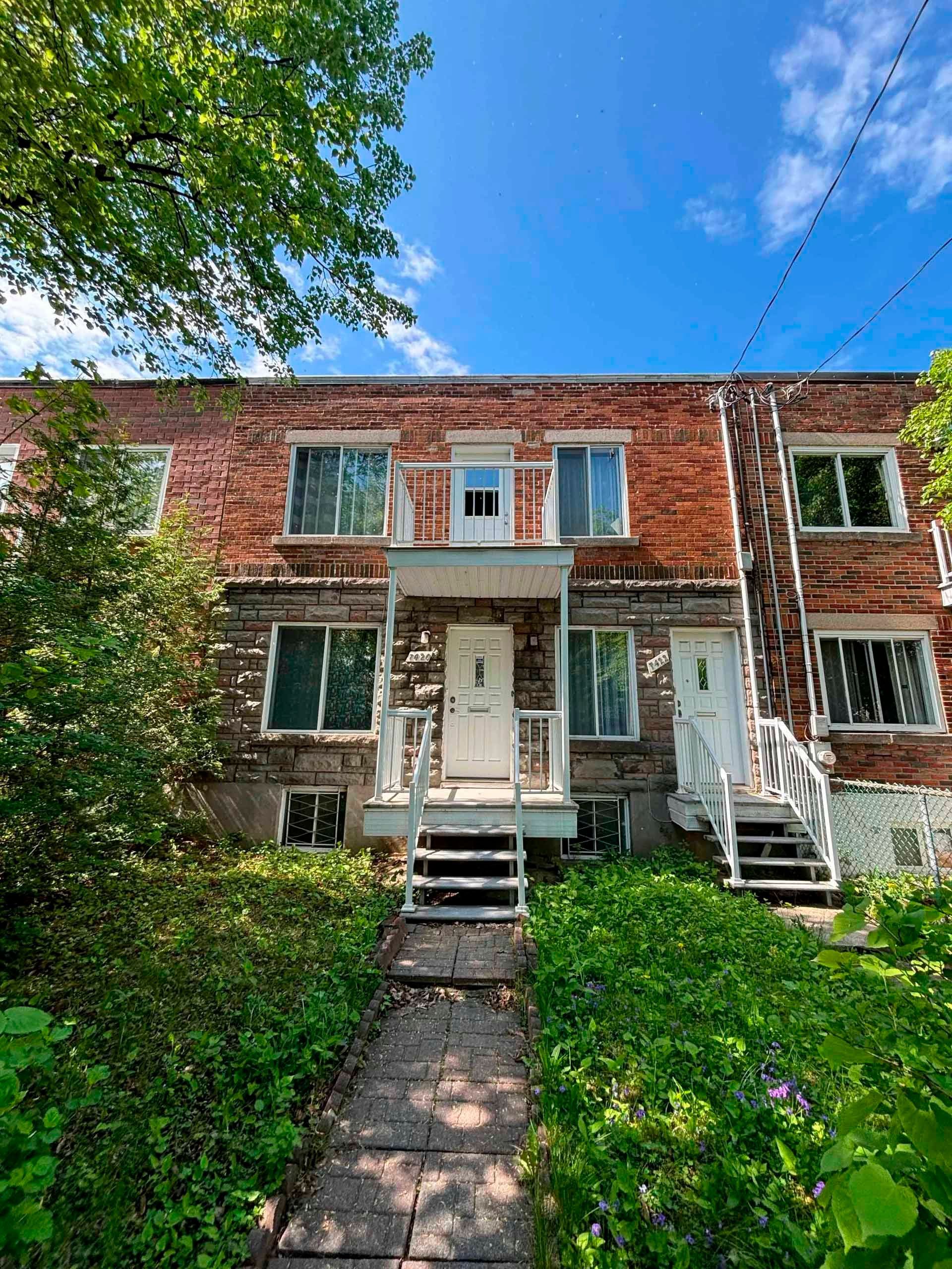 Immeuble de plusieurs logements en briques, avec portes et escaliers blancs, végétation envahissante à l'avant.