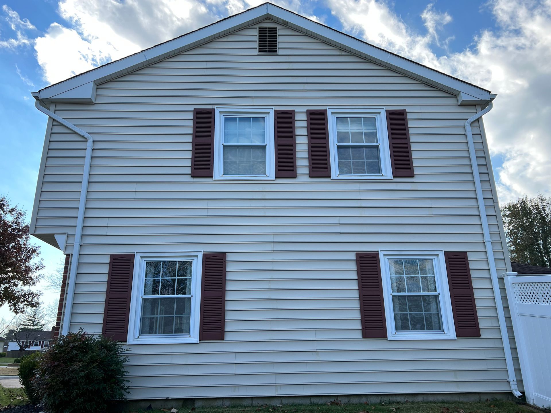 Two-story beige house with red shutters and white trim, under a partly cloudy sky.