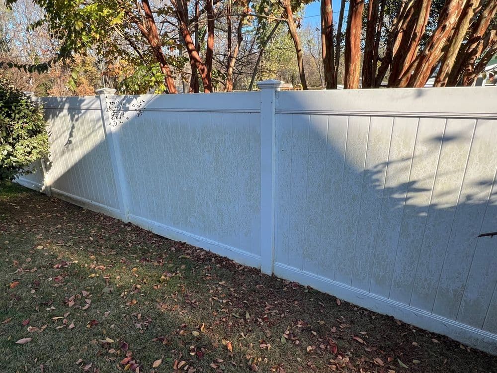 White vinyl fence in a grassy yard, speckled with discoloration. Sunlight casts shadows.