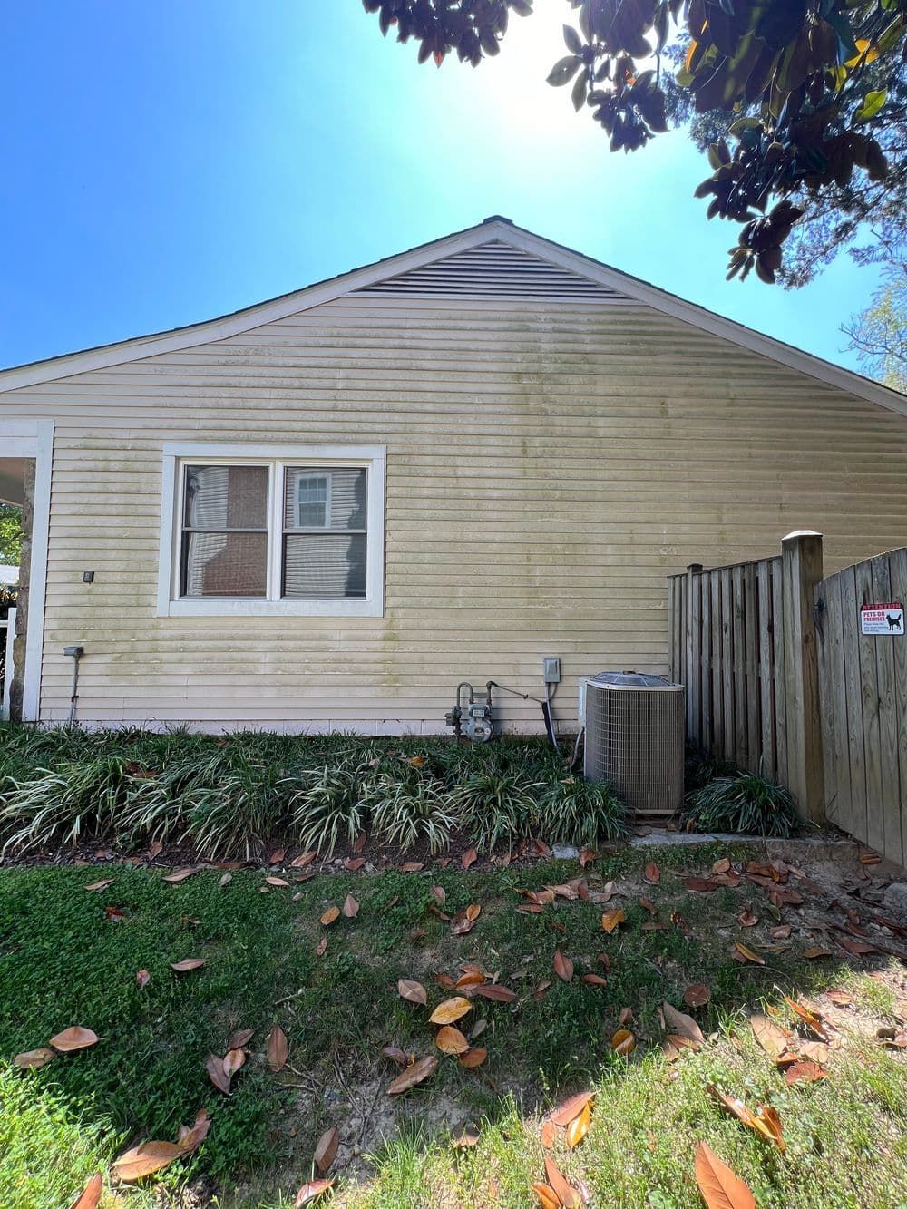 Yellow house with a window, air conditioner, and weathered siding. Fence on right, greenery below.
