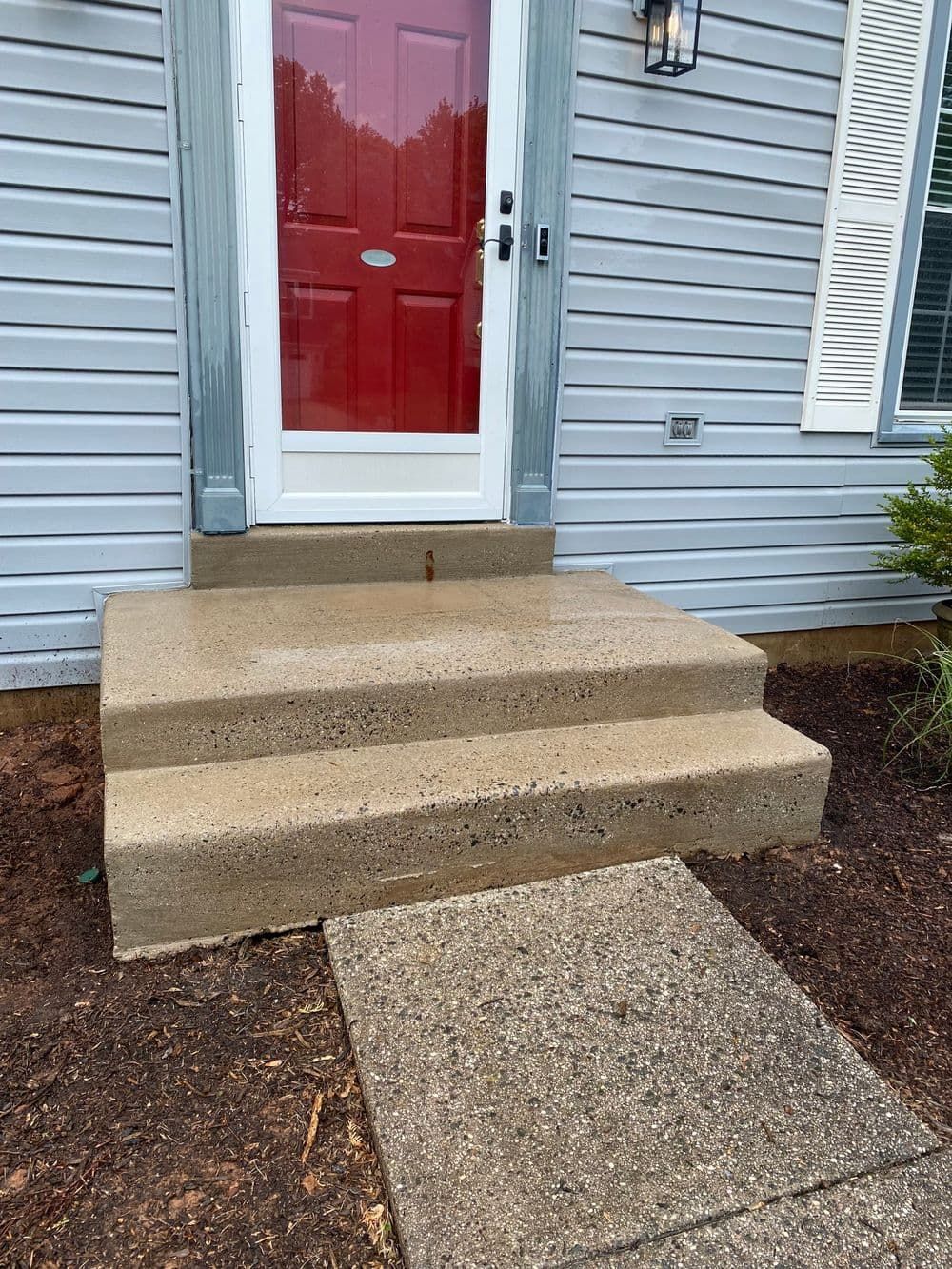 Concrete steps leading to a red door with a clear storm door. Gray siding and a concrete walkway.