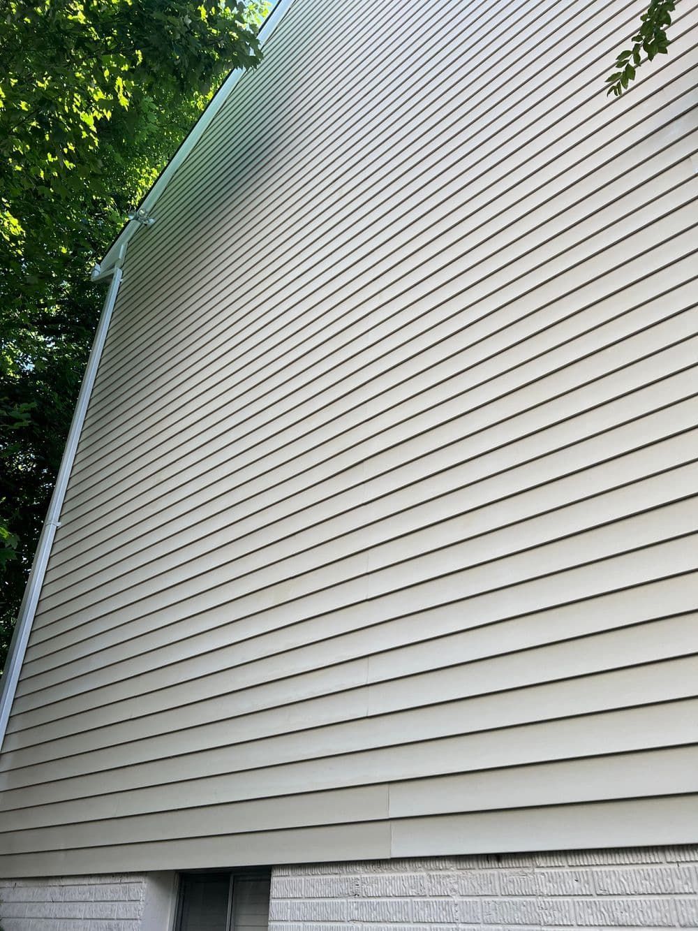 Tan siding on a building with a white trim and a small stone foundation. Green tree visible.