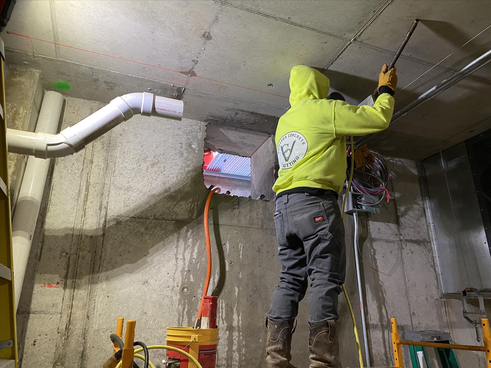 A man in a yellow hoodie is working on a ceiling in a basement.