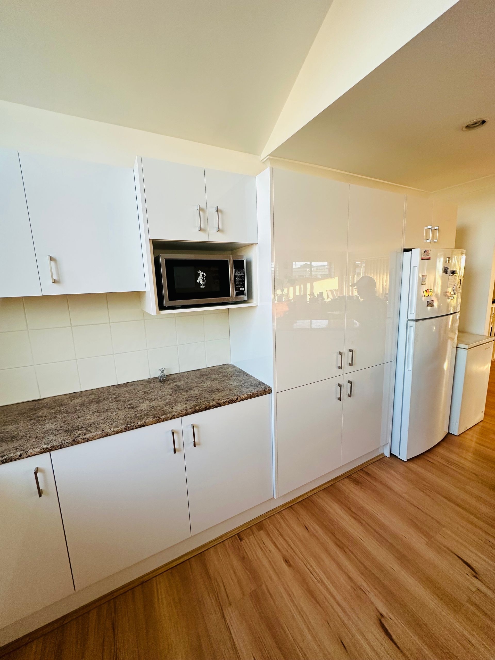 A Kitchen With Glossy White Cabinets, A Refrigerator And A Microwave — Nyon Shopfitters in Morisset, NSW
