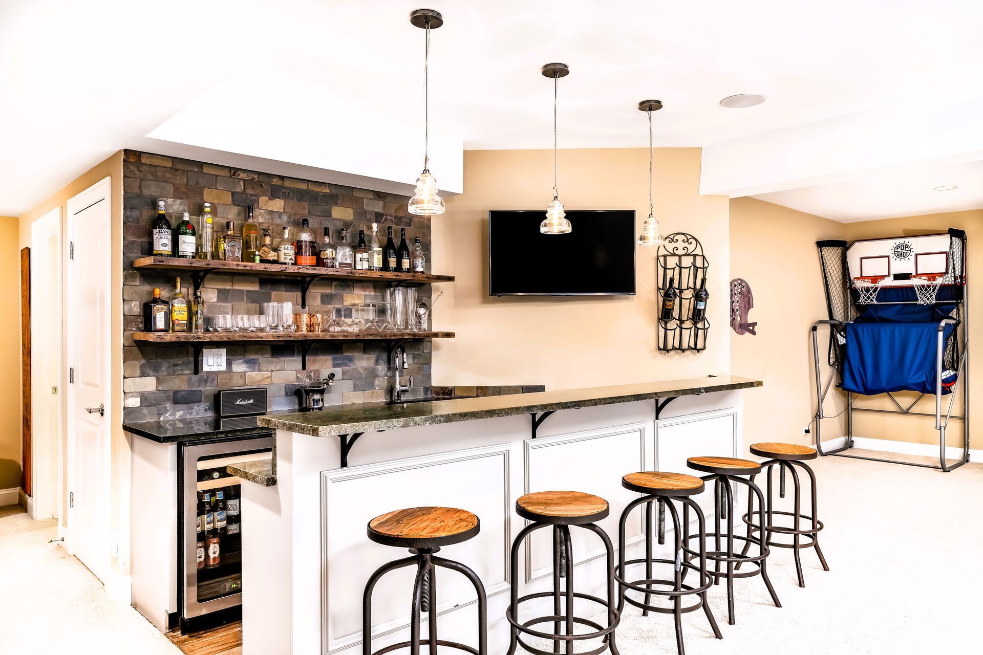 Modern basement bar with brick wall, granite countertop, wooden stools, TV, and basketball arcade game.