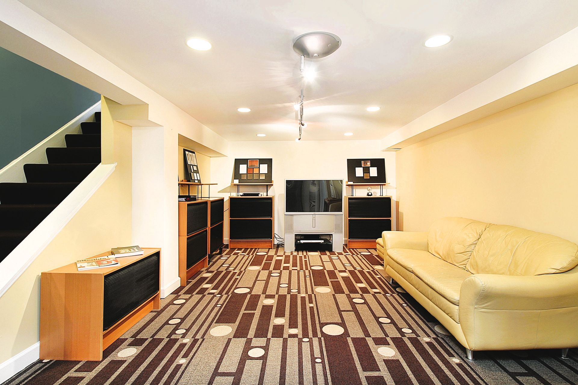 Modern basement with beige sofa, patterned carpet, organized shelves, and staircase, post-spring cleaning.