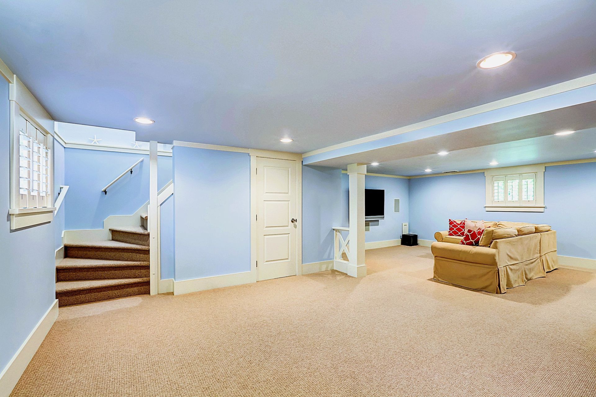 Light blue basement with beige carpet, red pillows, and staircase.
