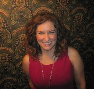 Woman with curly brown hair, red dress, smiling in front of patterned wallpaper.