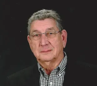 Older man in glasses, black jacket, patterned shirt; studio portrait, neutral expression.