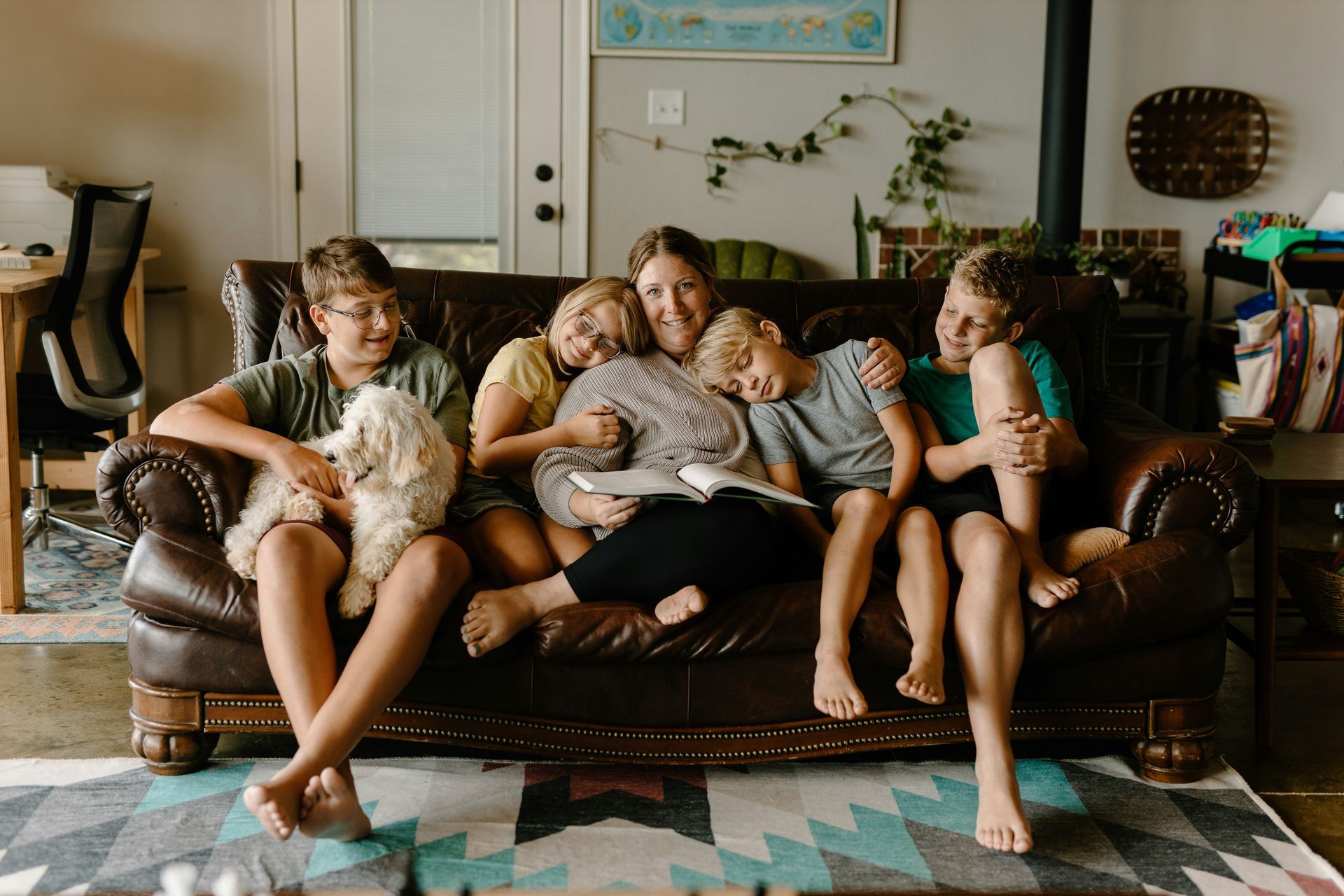 Family of six cuddling on a brown leather couch; a mother reading a book with her children, some hugging her.