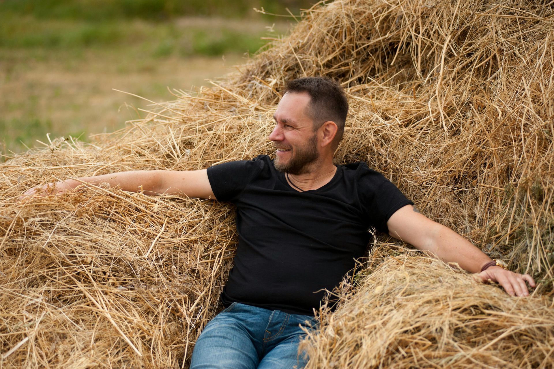 Man in black shirt and jeans smiling, leaning against a hay bale in a field.