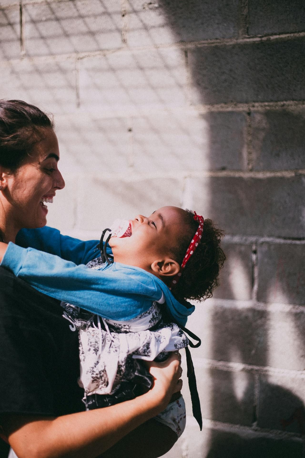 Woman holding a laughing child outdoors; brick wall in the background, sunny with shadow.