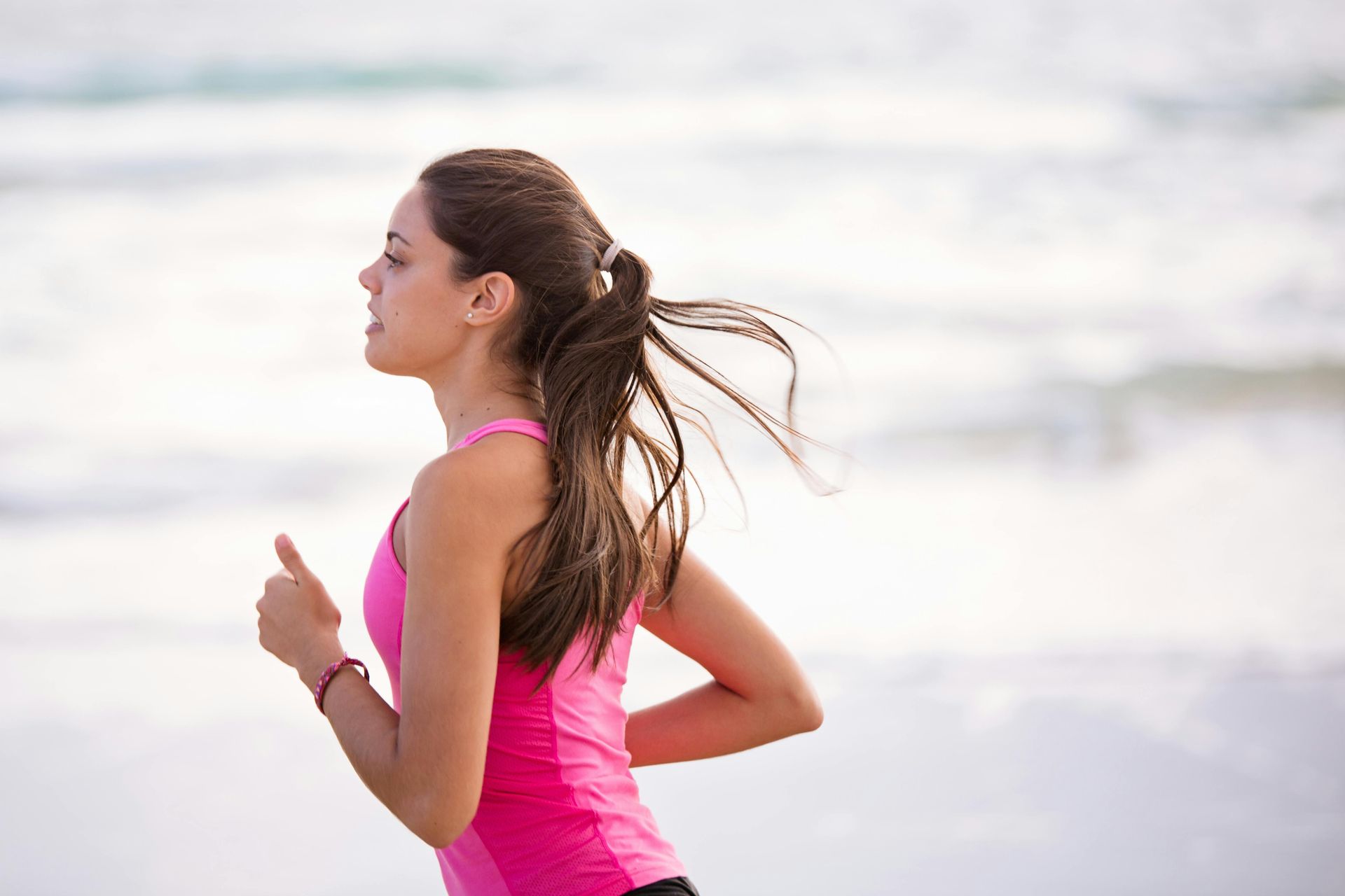 Woman in pink tank top running on a beach, hair in ponytail, ocean in background.