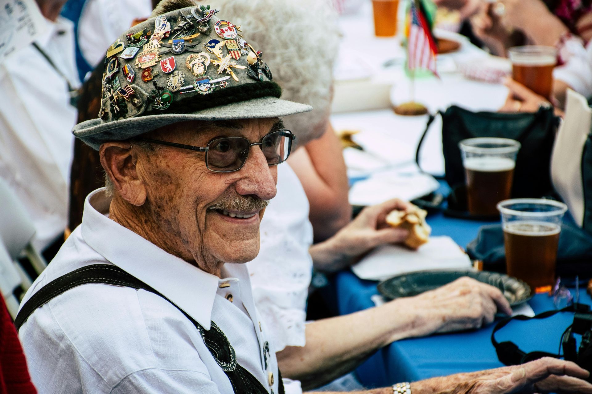 Elderly man wearing a hat covered in pins smiles while sitting at a table with beer.