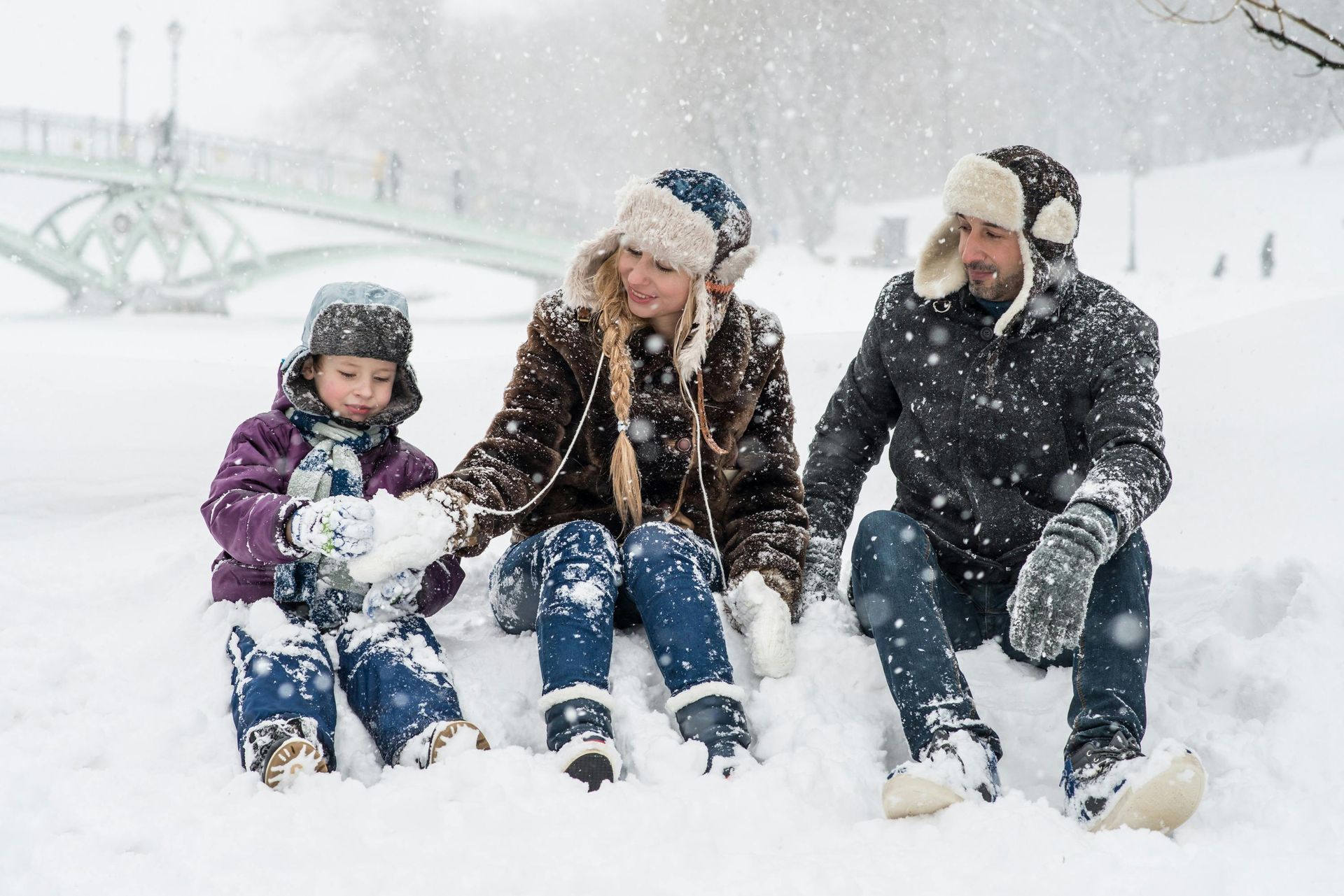 Family of three sits in snow, building snowballs. Snowy bridge in background.