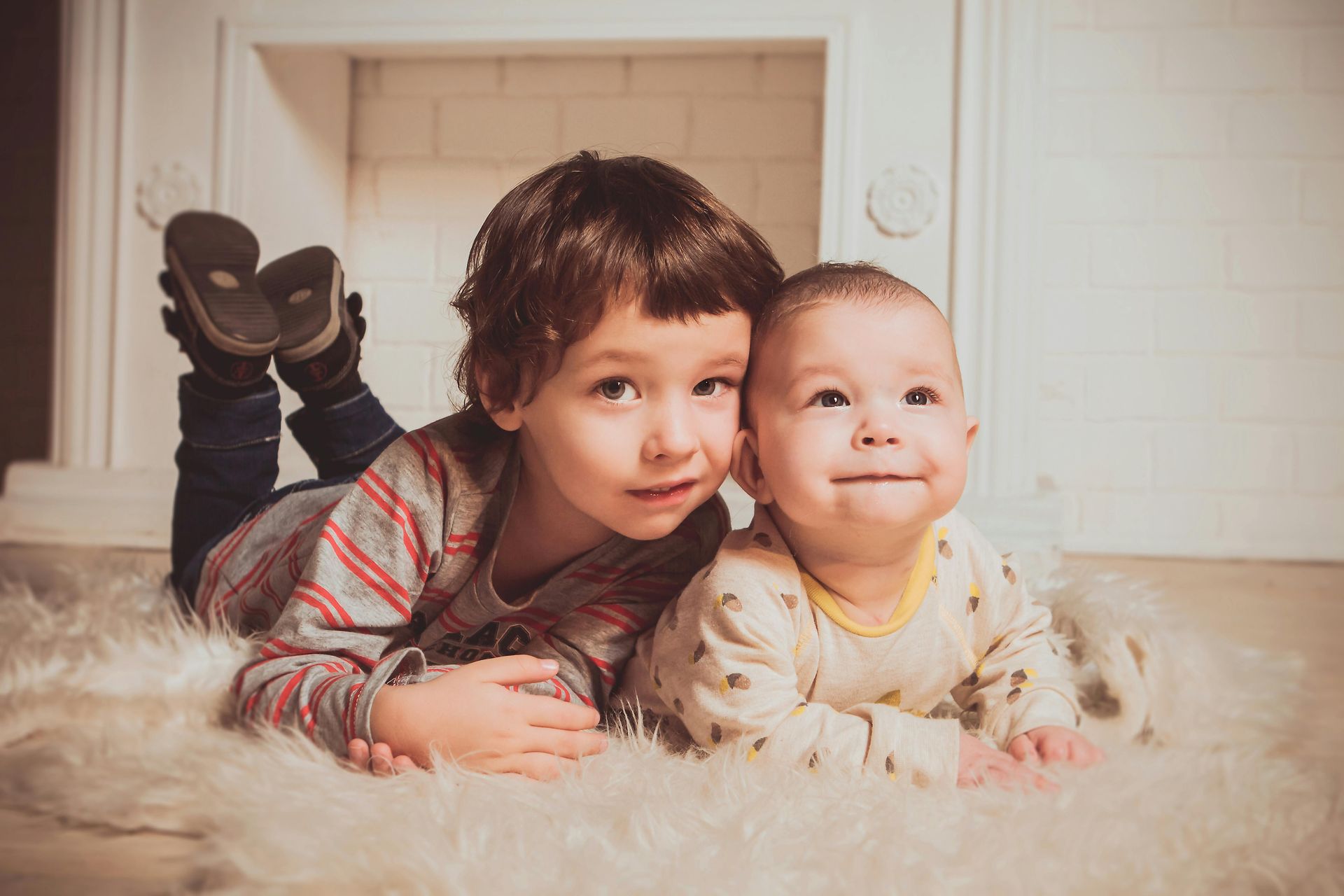 Two young children lying on a fluffy rug, one with a striped shirt, the other a baby in a onesie, smiling.