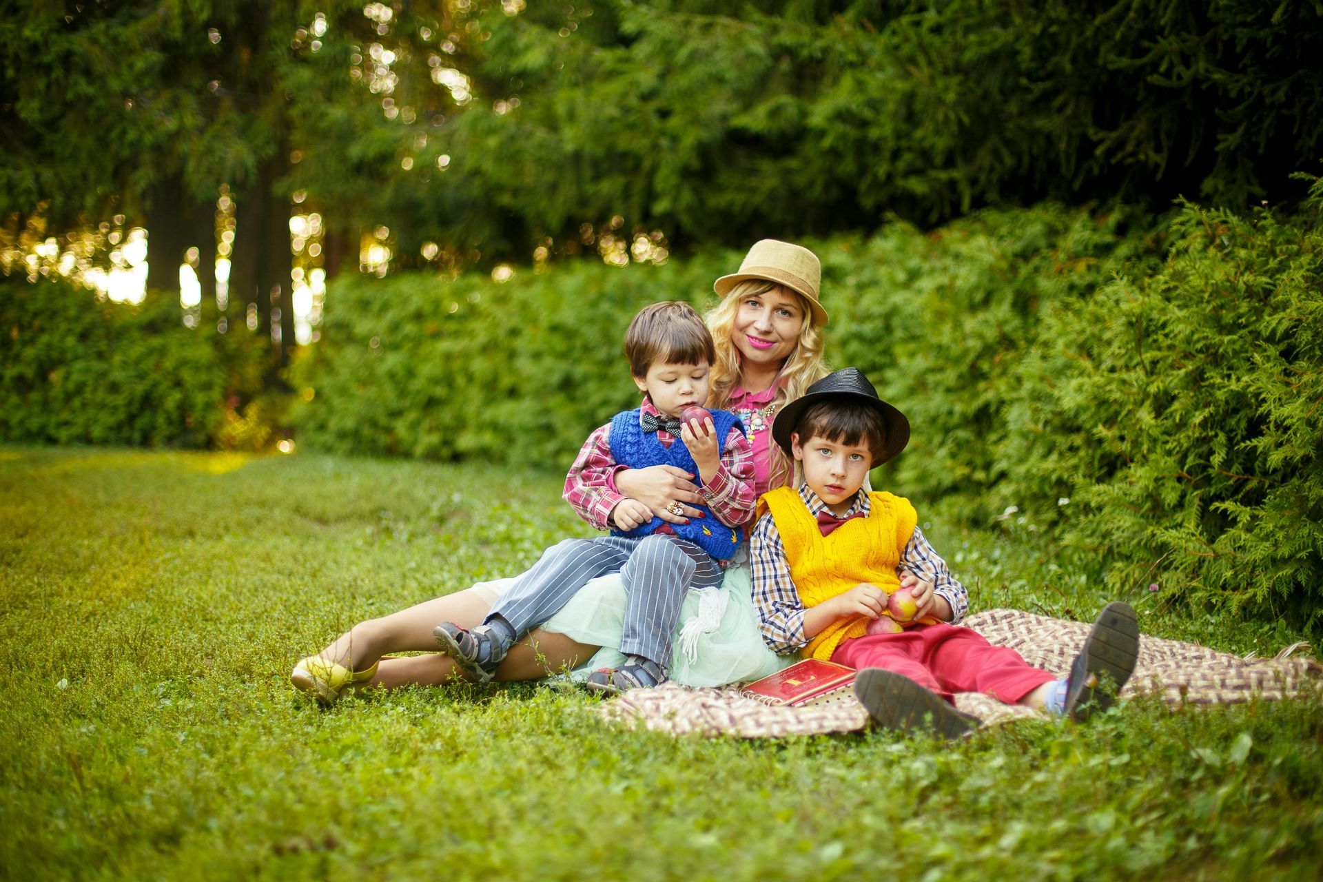 Woman and two children sit on a blanket in a park. They're smiling.
