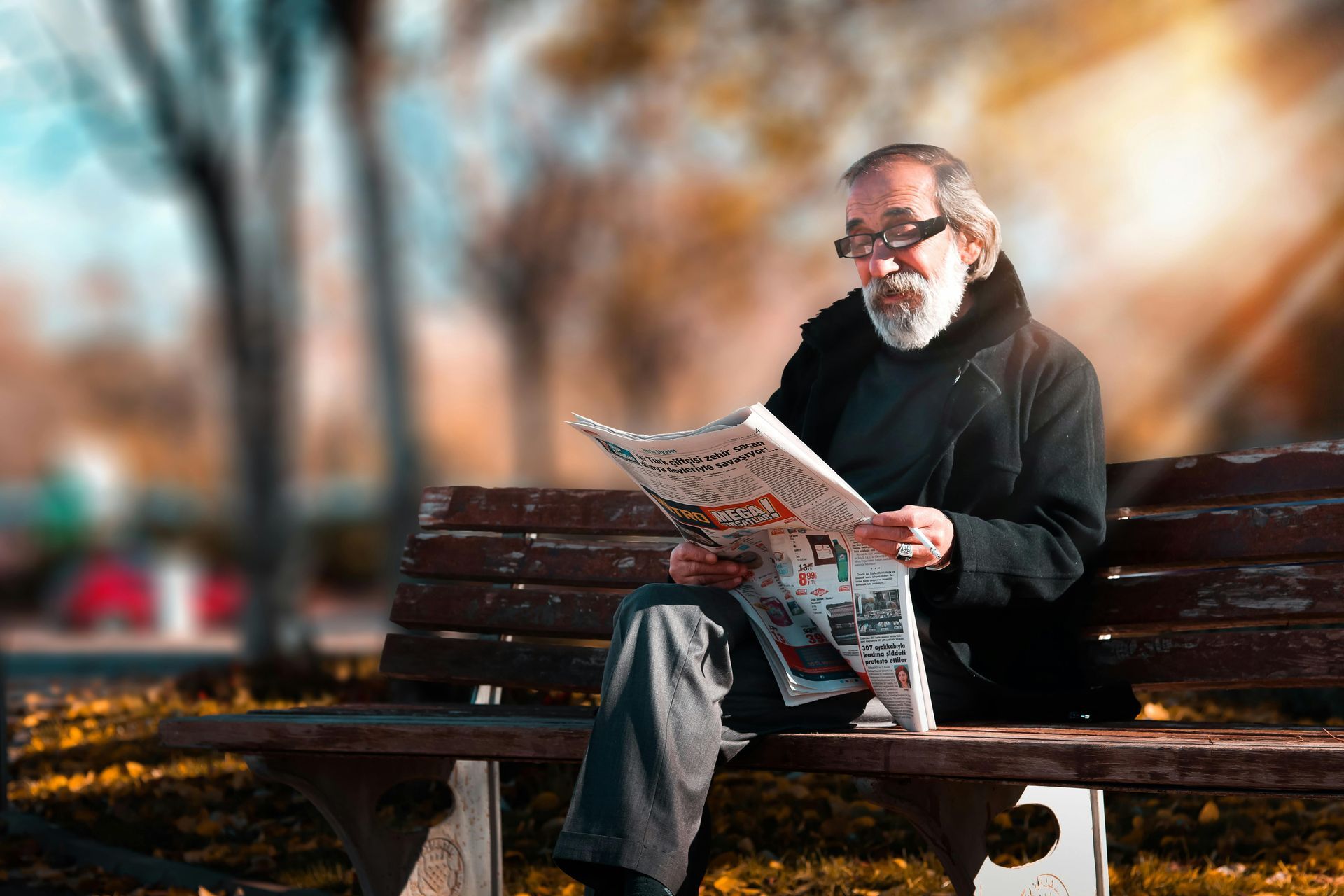 Elderly man with glasses reads newspaper on a park bench; autumn leaves and sunlight in the background.