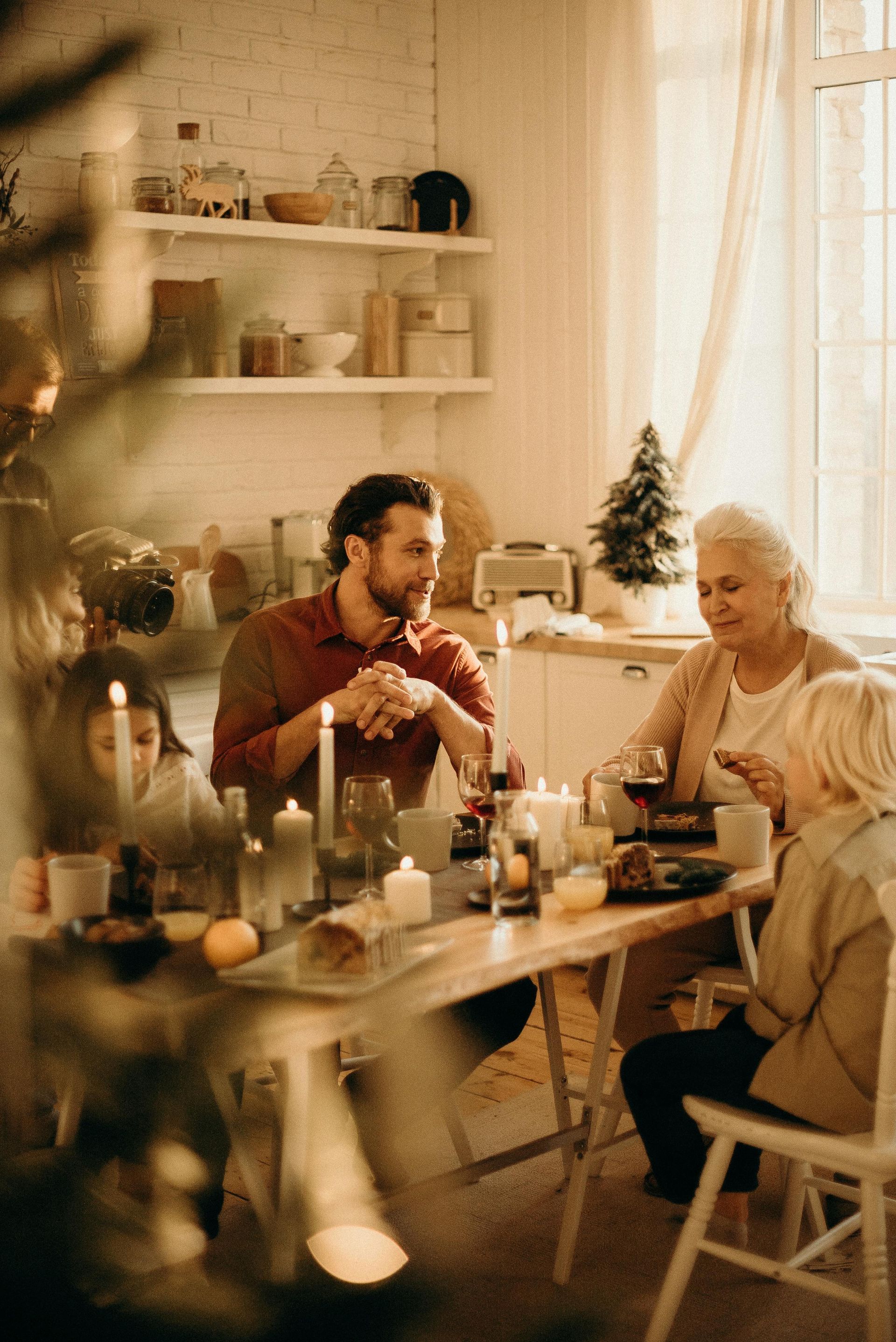 Family at a dining table, lit by candles; man talking, grandmother smiling, Christmas tree in background.