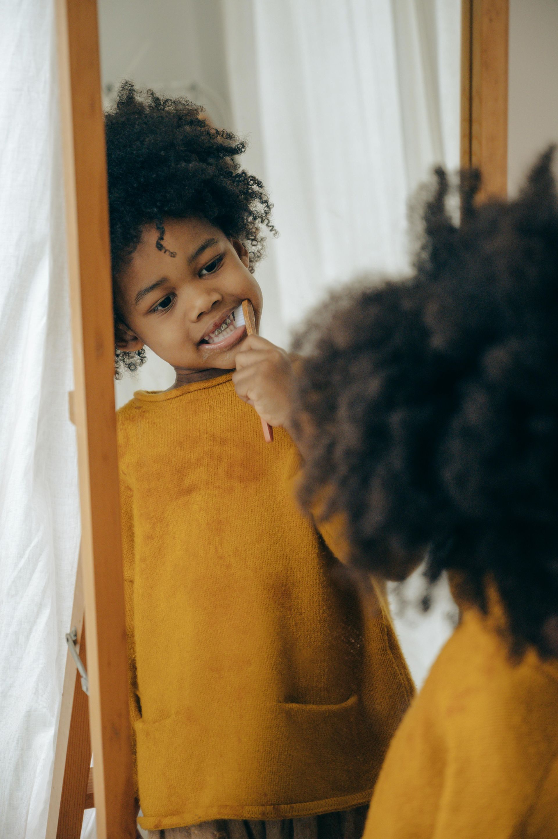 Black child in yellow shirt brushing teeth in front of a mirror.