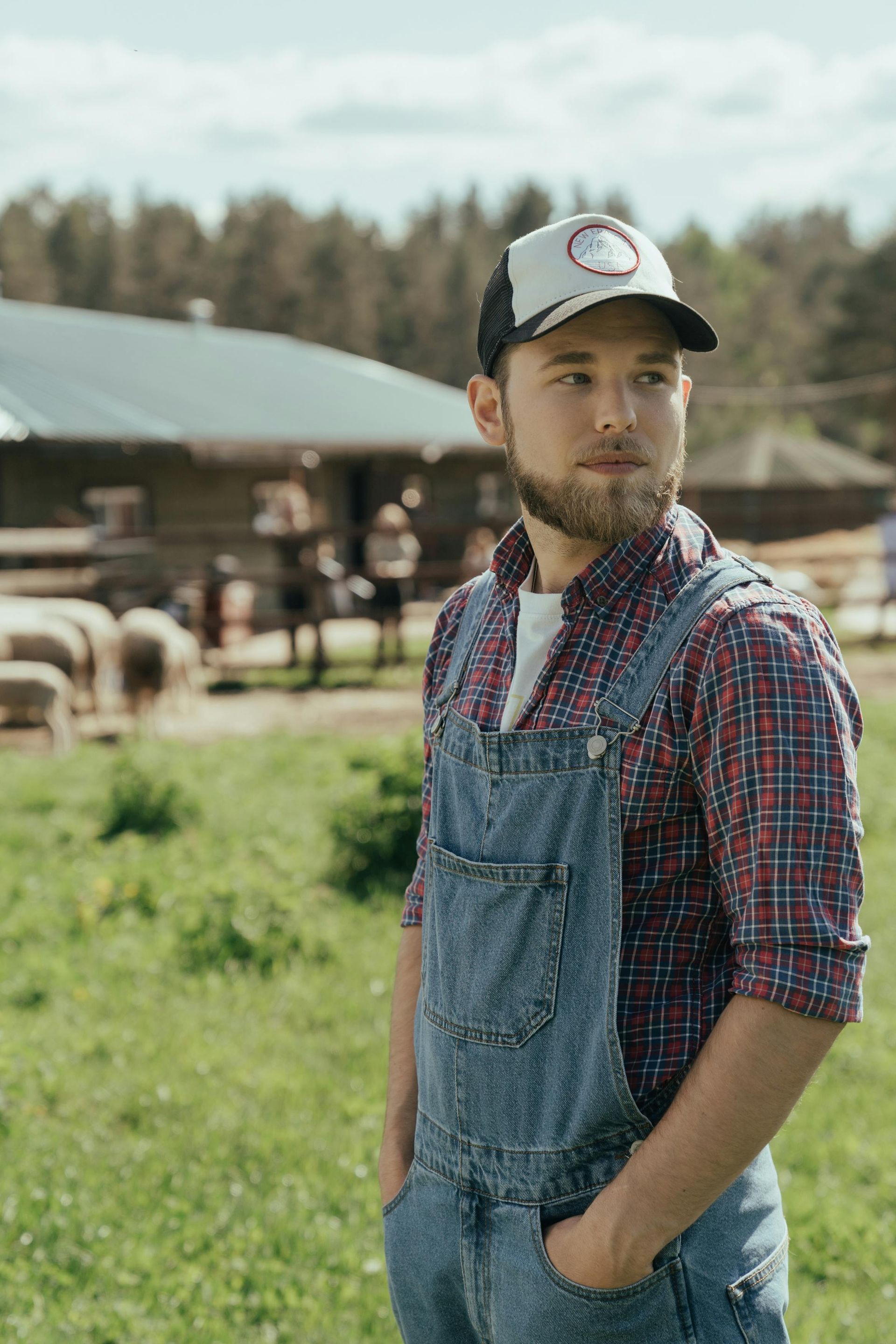 Man in overalls and plaid shirt with hands in pockets, on a farm, looking away.