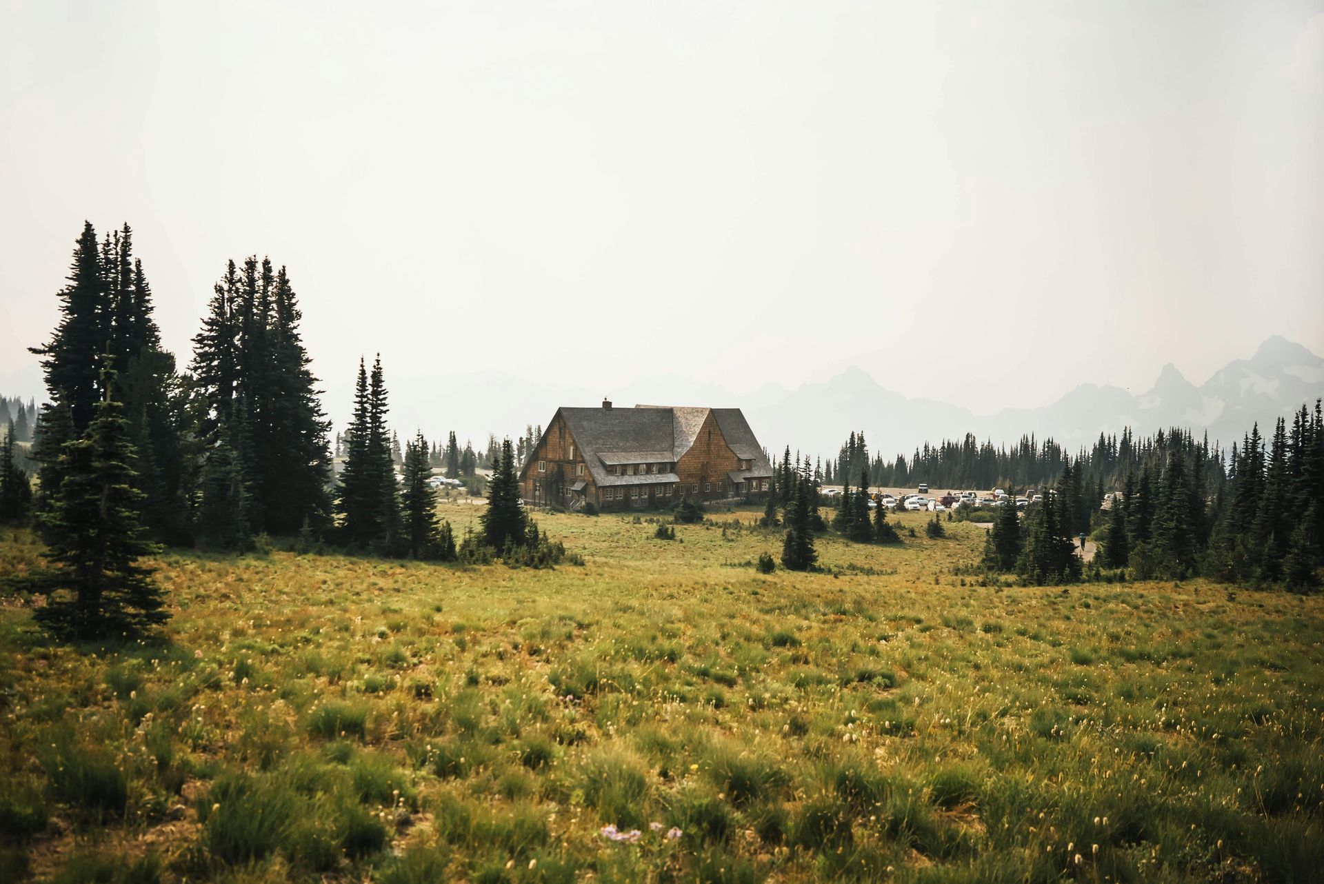 A large, rustic lodge sits in a grassy meadow, surrounded by evergreen trees, mountains in the distance under a hazy sky.