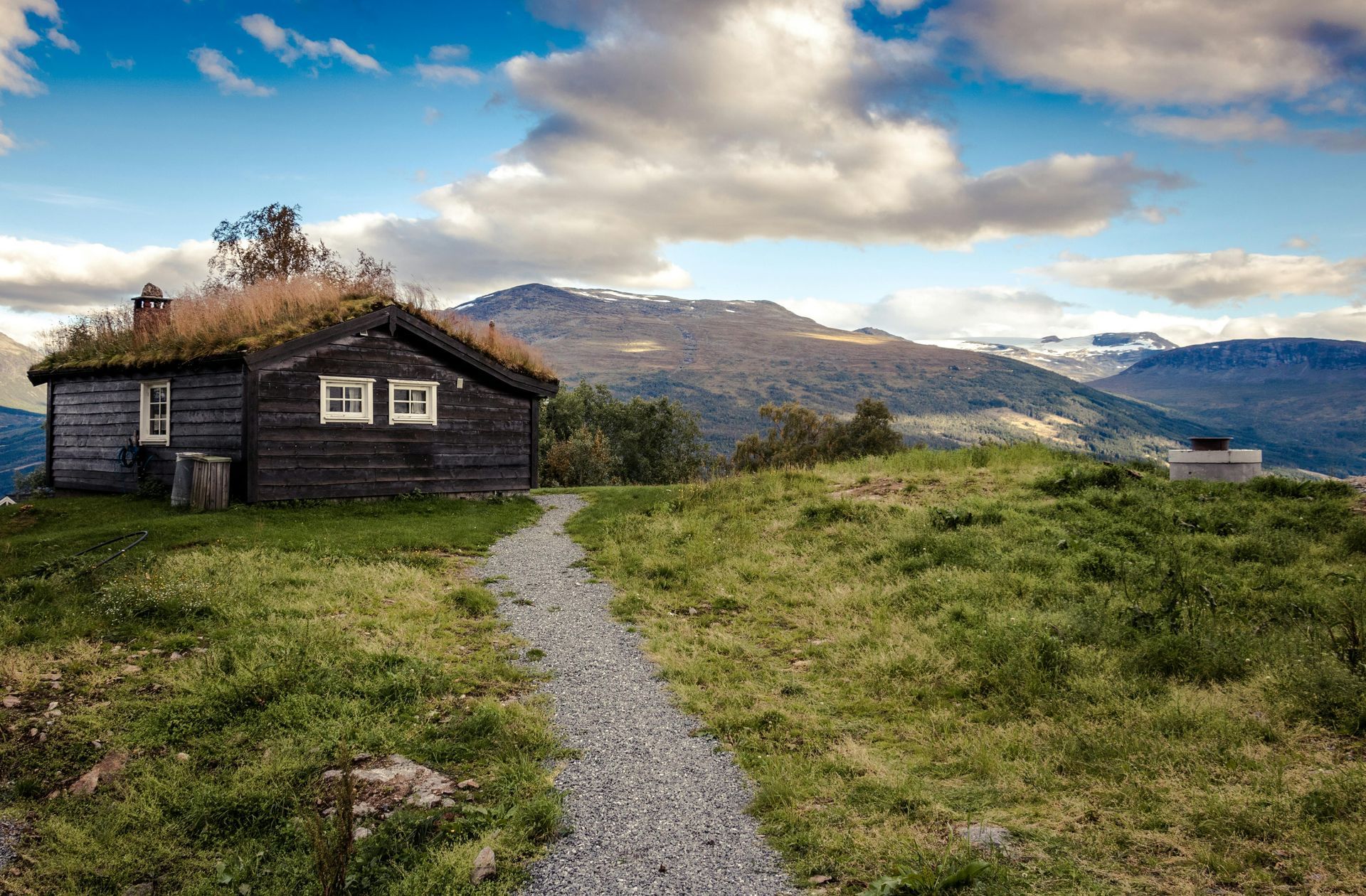 A rustic cabin with a grass roof sits atop a grassy hill, overlooking a mountain range under a cloudy sky.