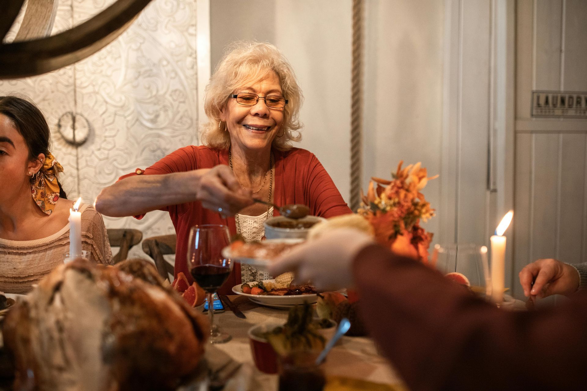 Elderly woman smiling, serving food at Thanksgiving table with family. Candles, turkey, flowers.