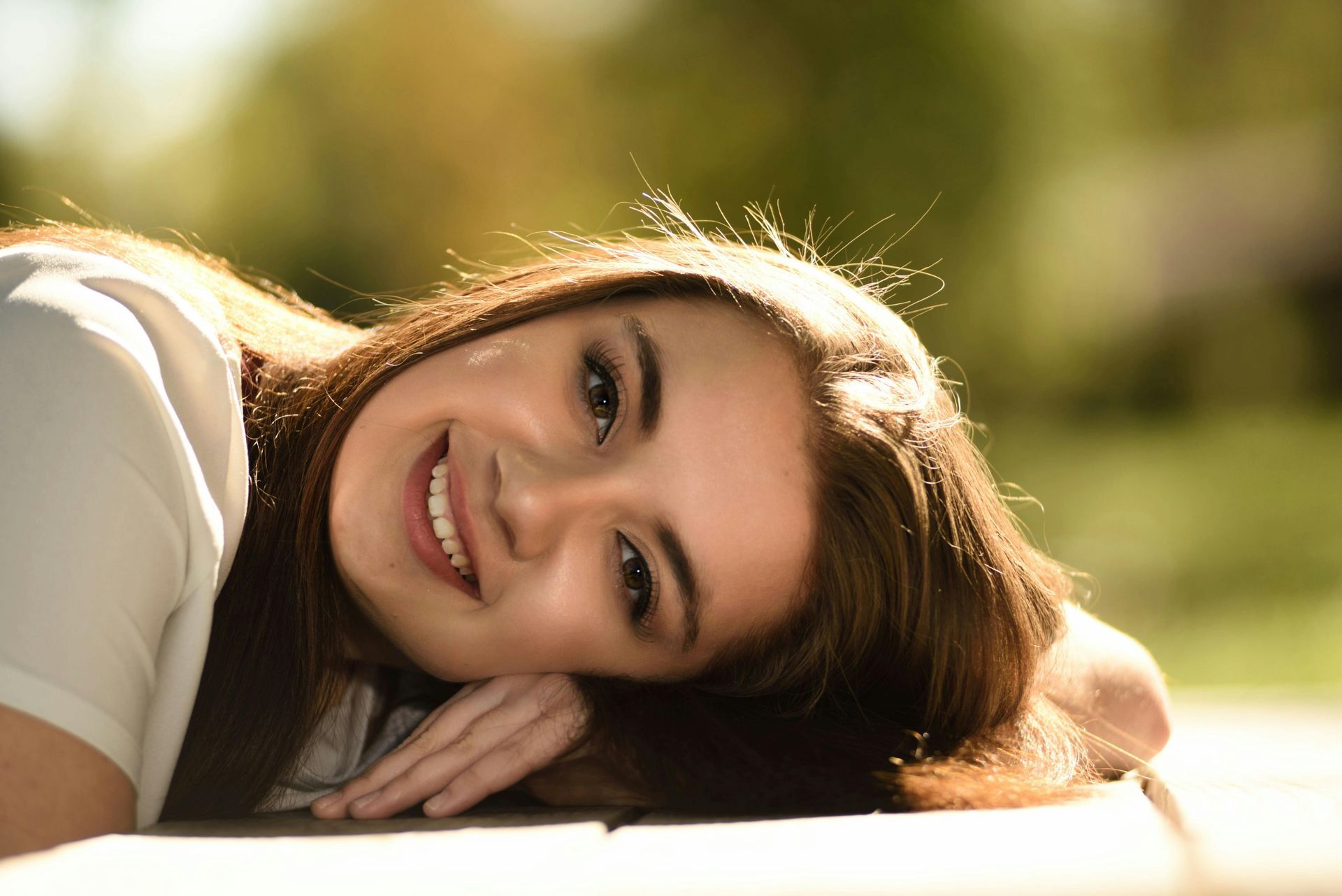 Woman smiles, leaning on a wooden surface outside, with brown hair and sunlight.