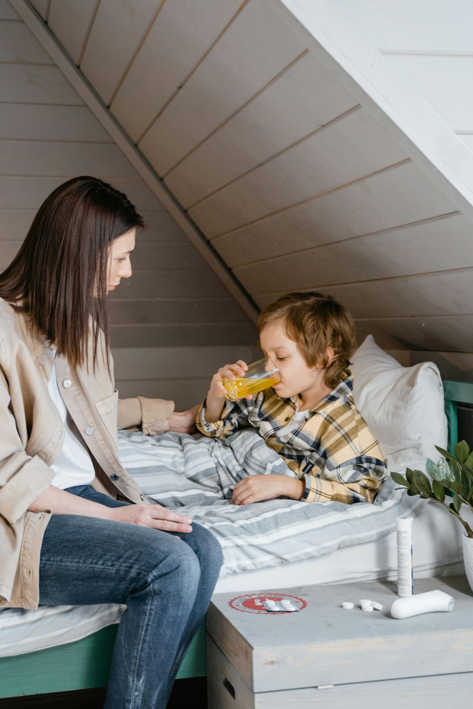 A woman sits with a child in bed, giving him orange juice. A box of medicine and a thermometer are on the bedside table.
