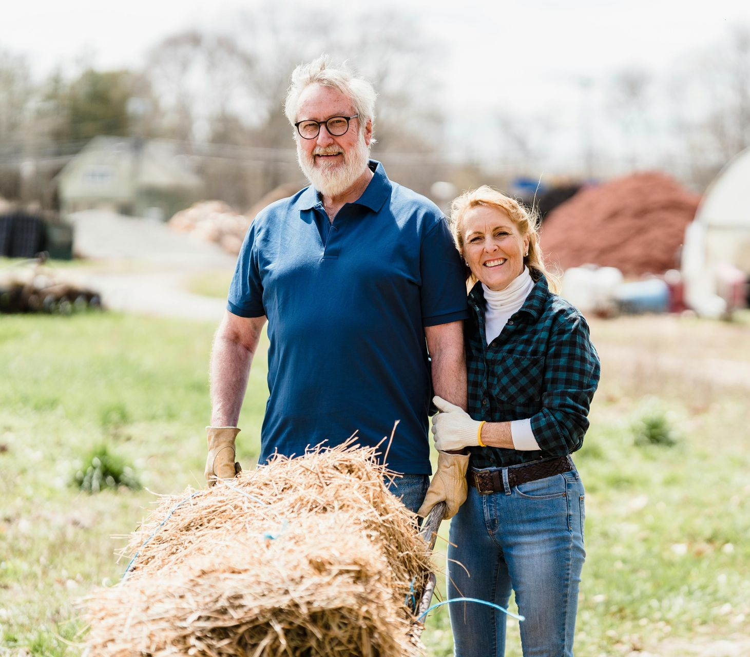 Man and woman with straw in field, smiling. Man wears blue shirt, glasses, woman wears flannel and gloves.