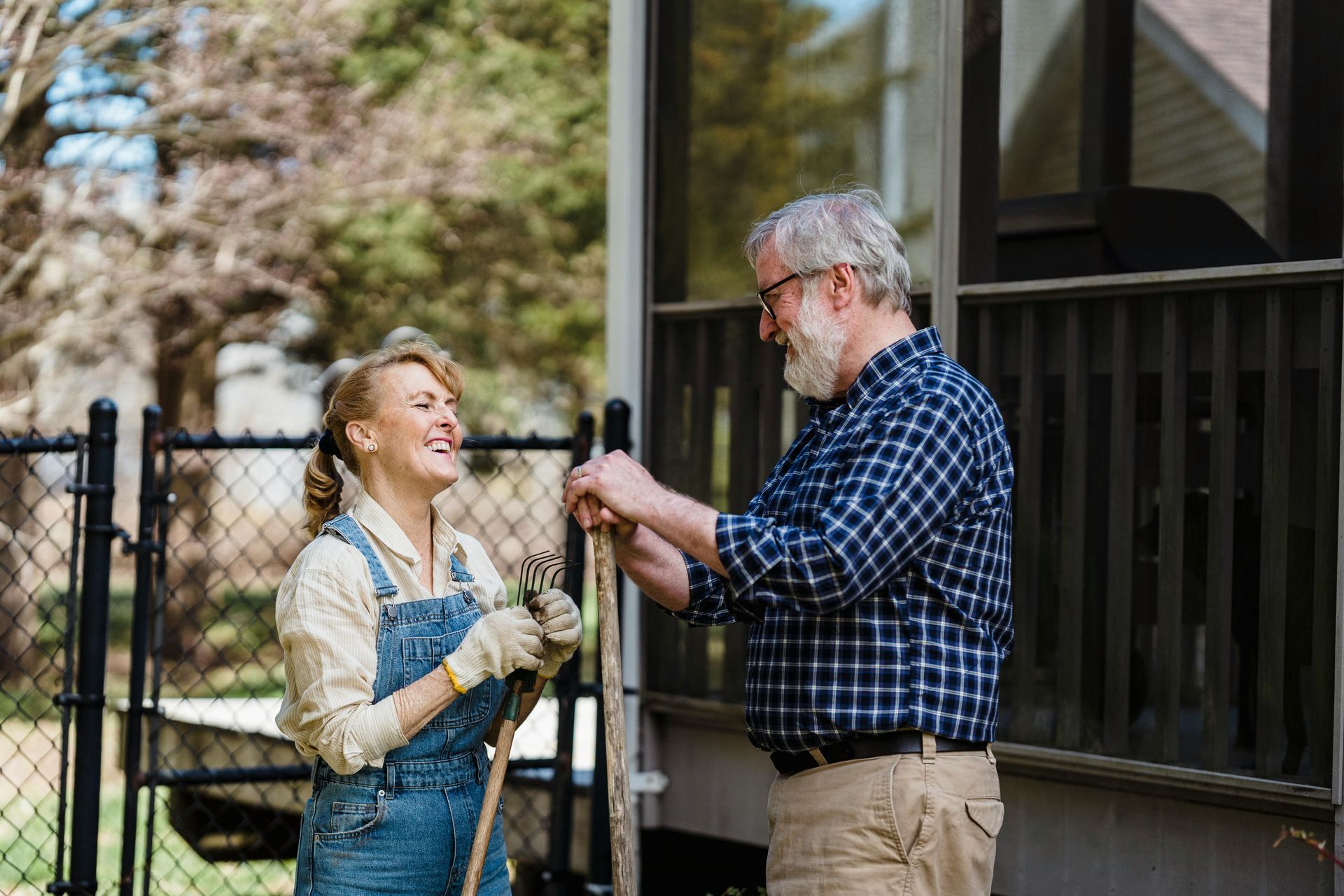 Woman and man gardening together, laughing. She wears overalls, he a plaid shirt. Outdoors by a house.