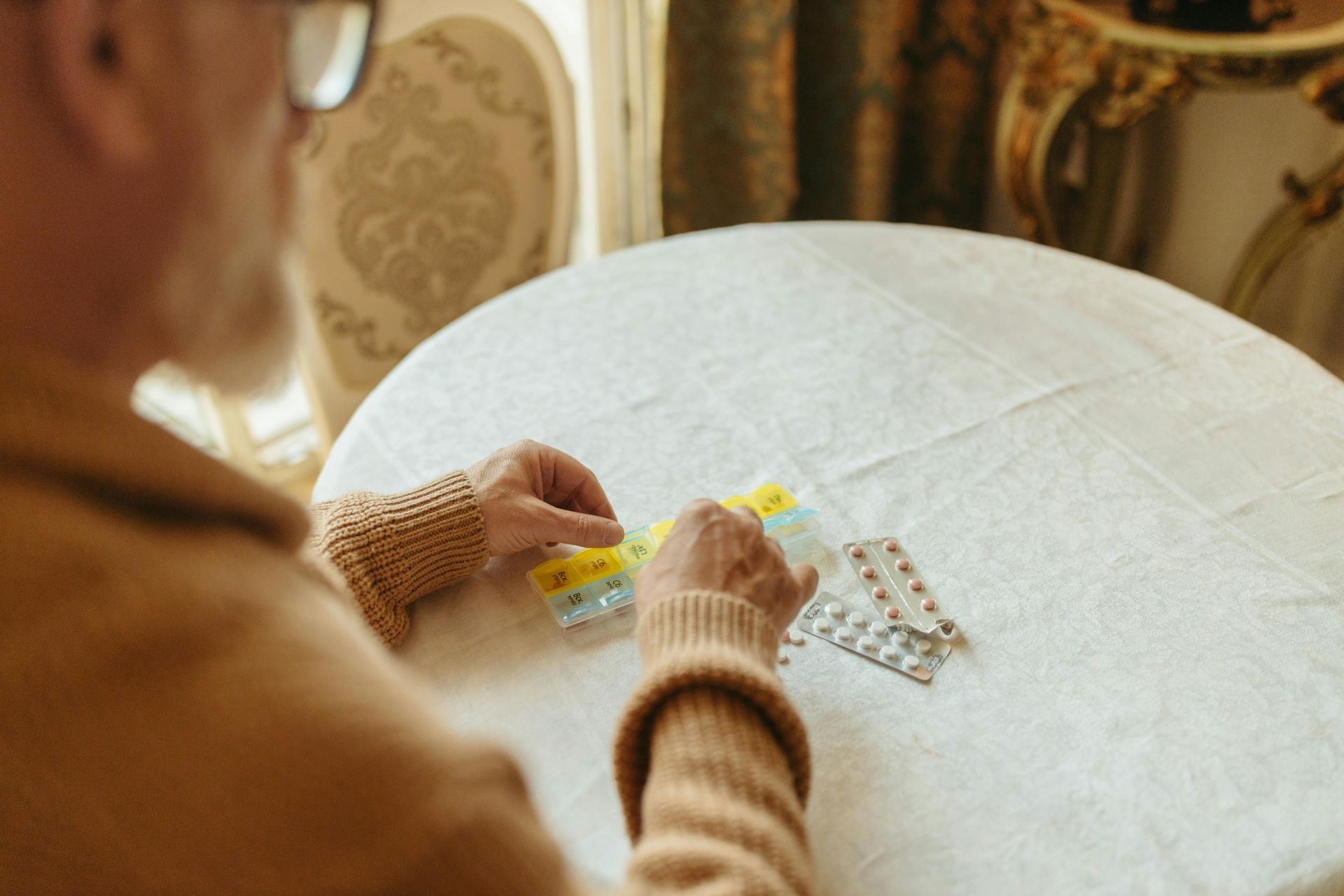 An older person with glasses sorts medication blister packs on a white tablecloth.